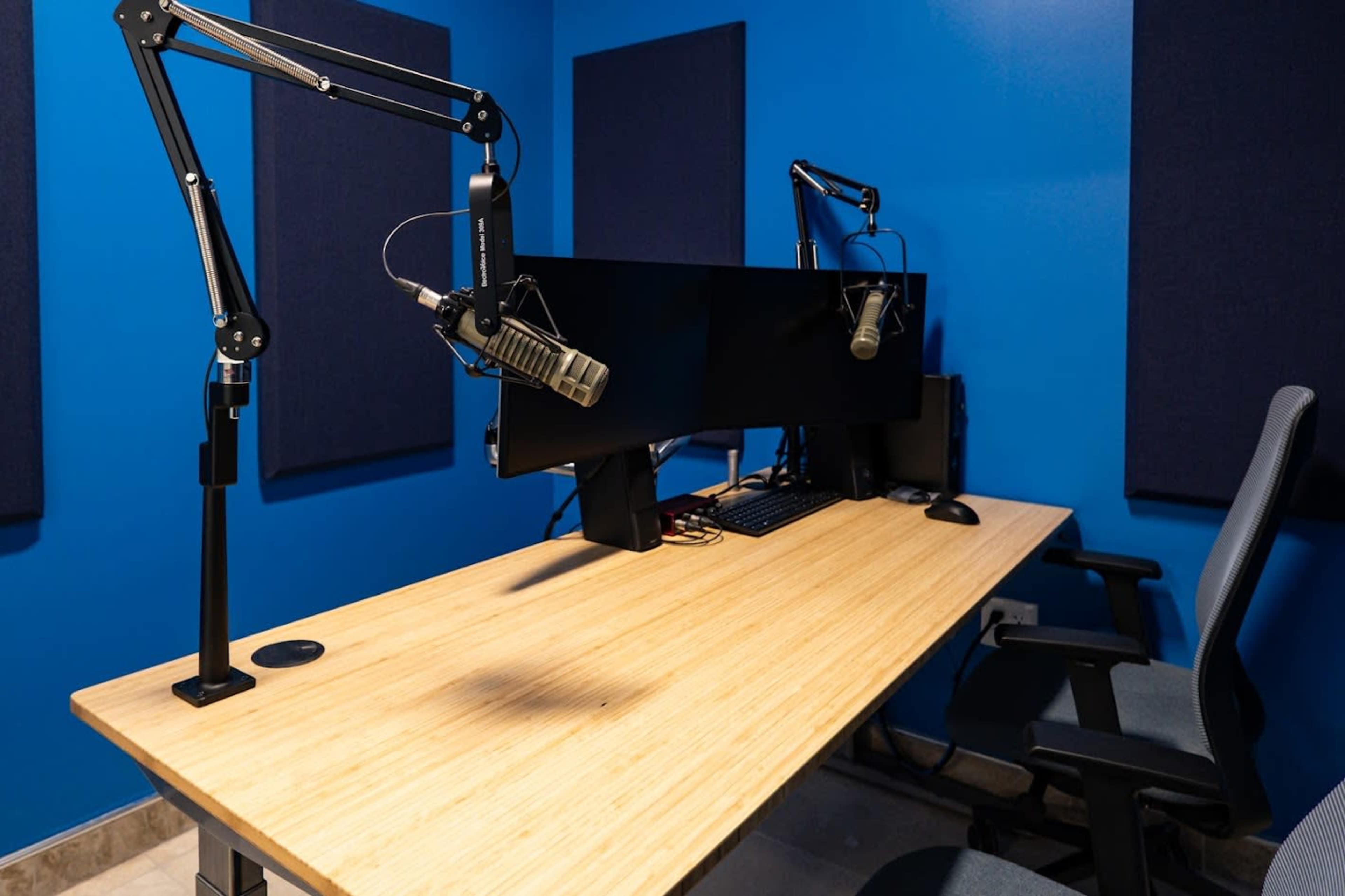 The image shows a modern podcast studio featuring a wooden table, two adjustable microphones, a dual-monitor setup, and soundproofed blue walls.