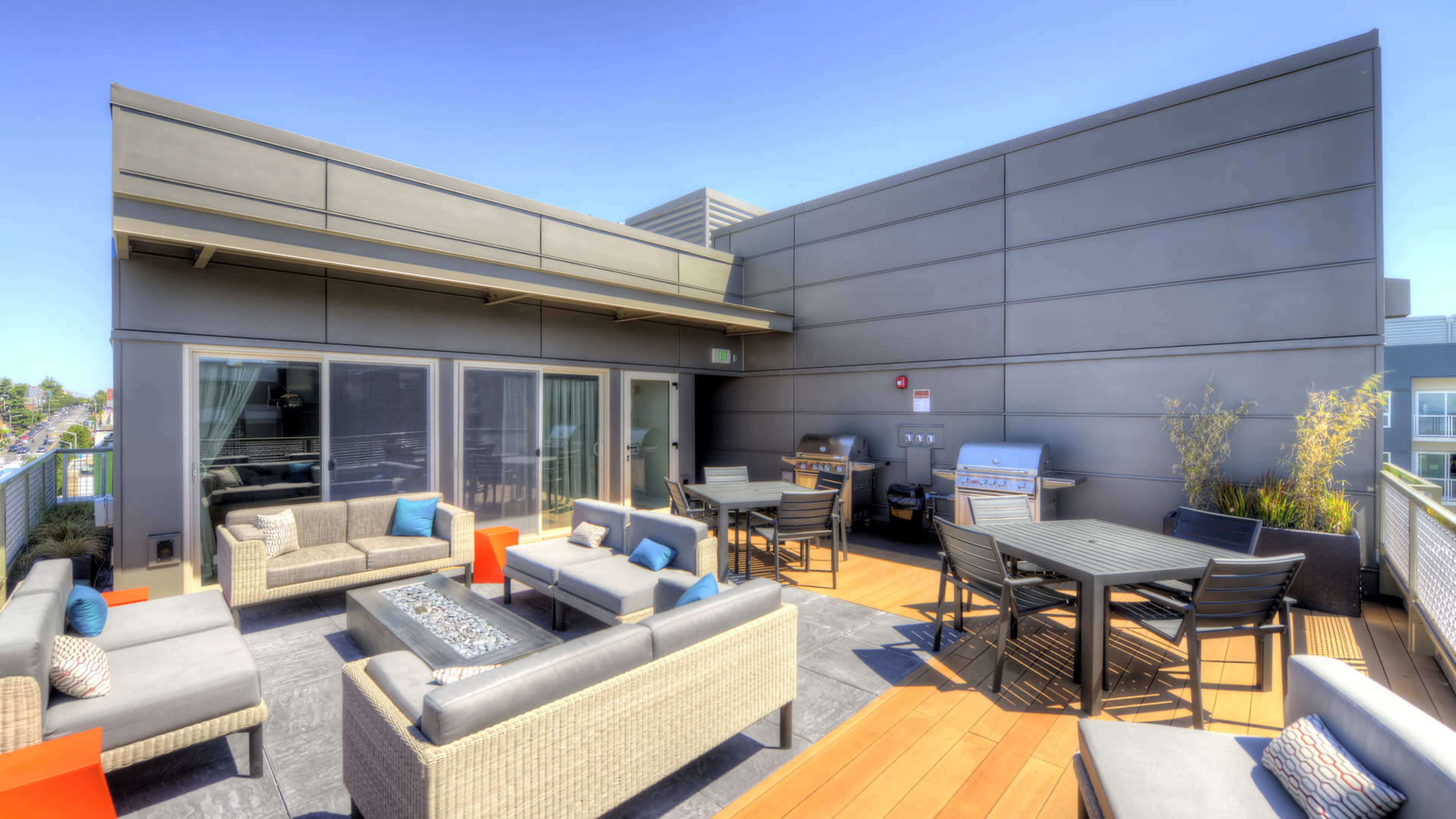 The rooftop terrace features lounge seating, a dining area, and a barbecue setup with a clear blue sky above.