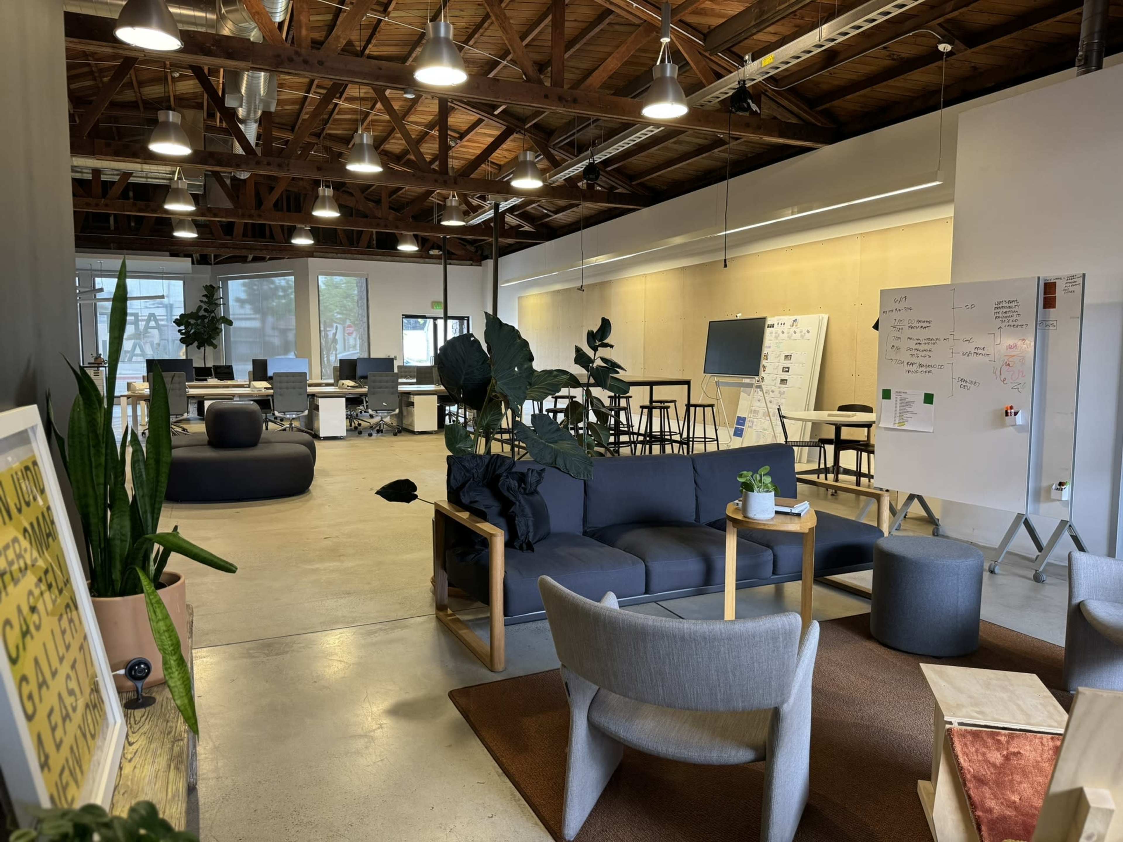 The image shows a modern office space with seating areas, meeting tables, and whiteboards, all under a wooden ceiling.