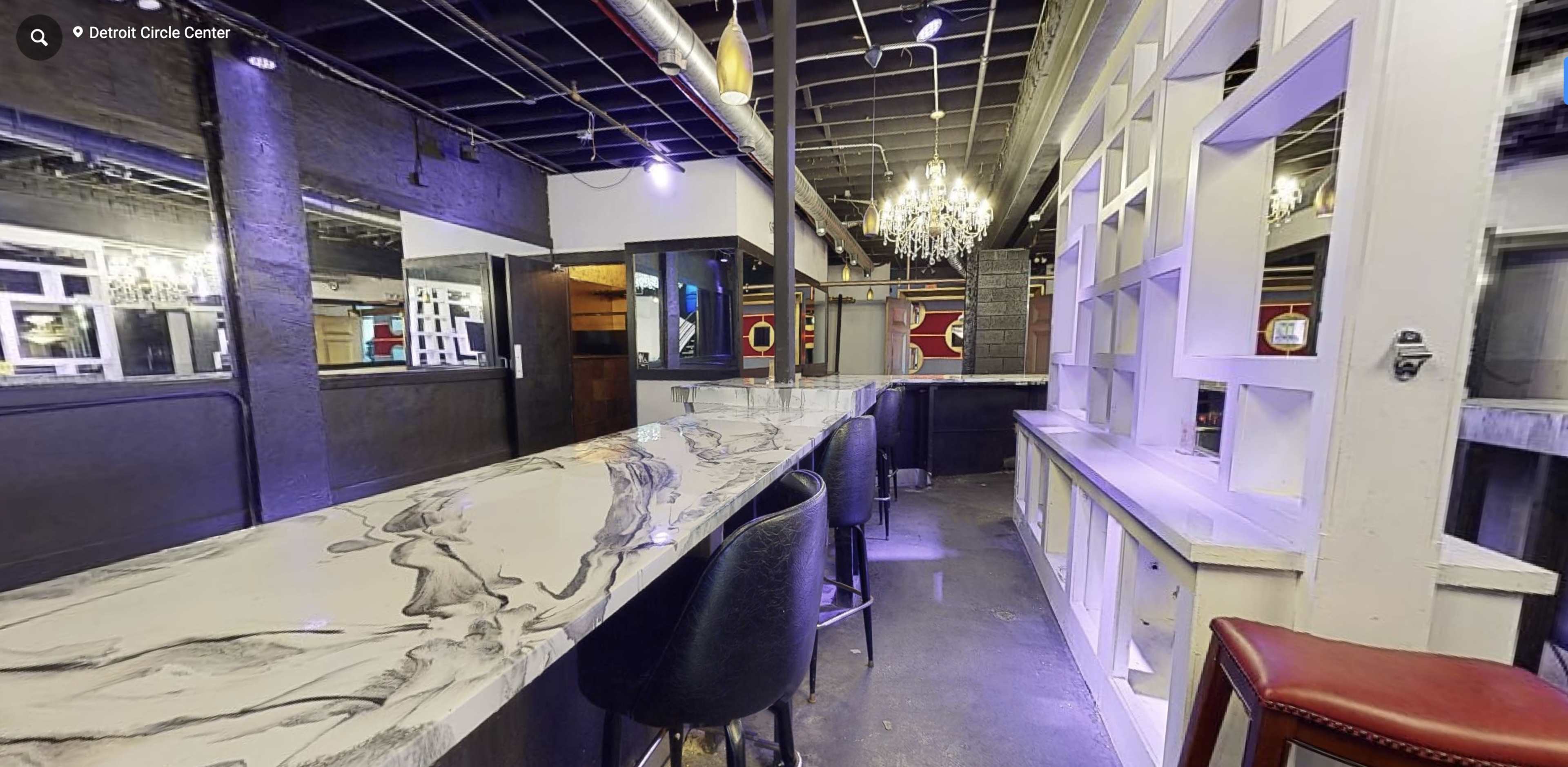 A modern bar area with a long, decorated countertop, high stools, and a chandelier overhead in a dimly lit space.
