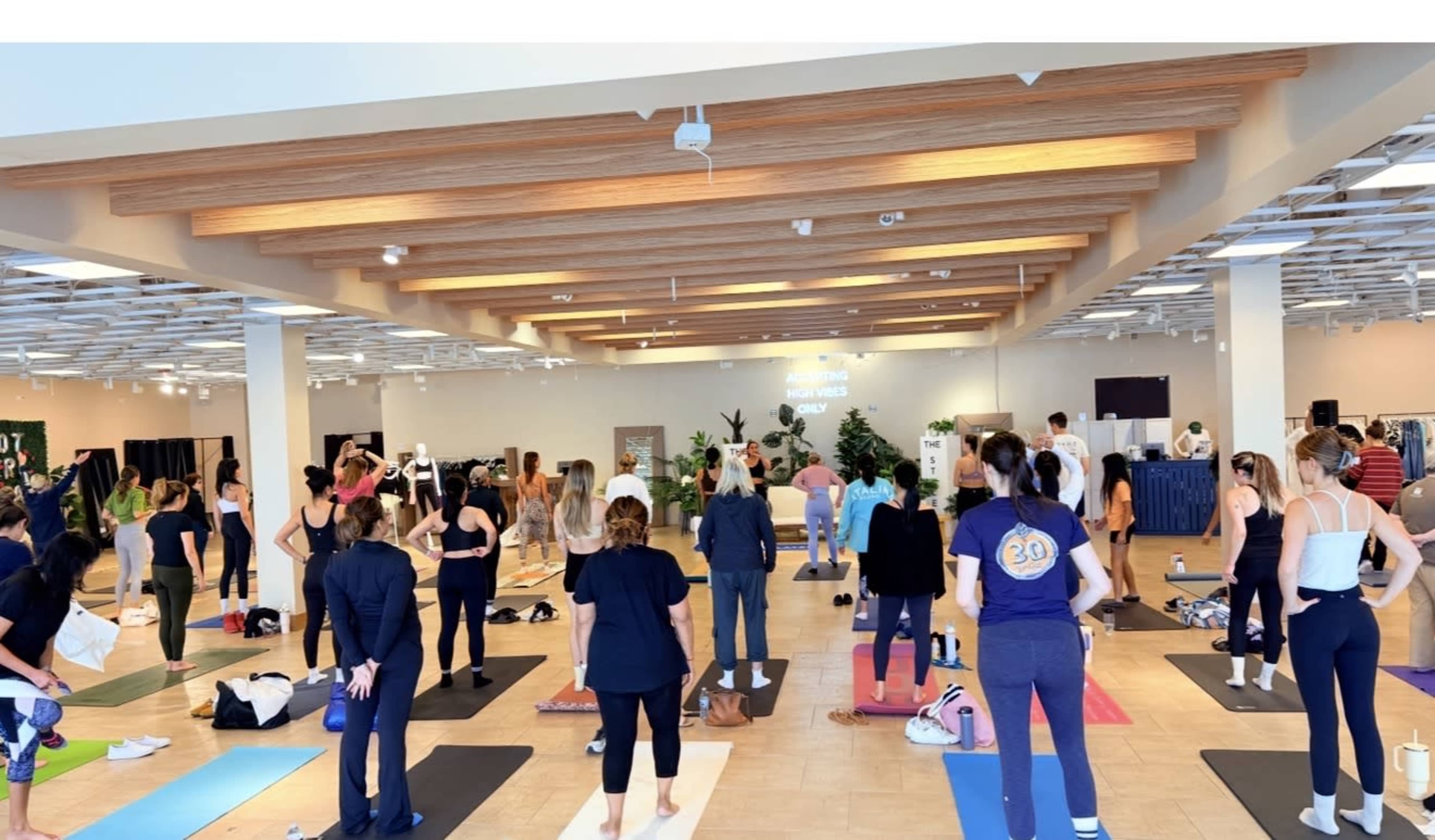 A group of people are participating in a yoga class in a spacious studio with wooden beams and mats arranged on the floor.