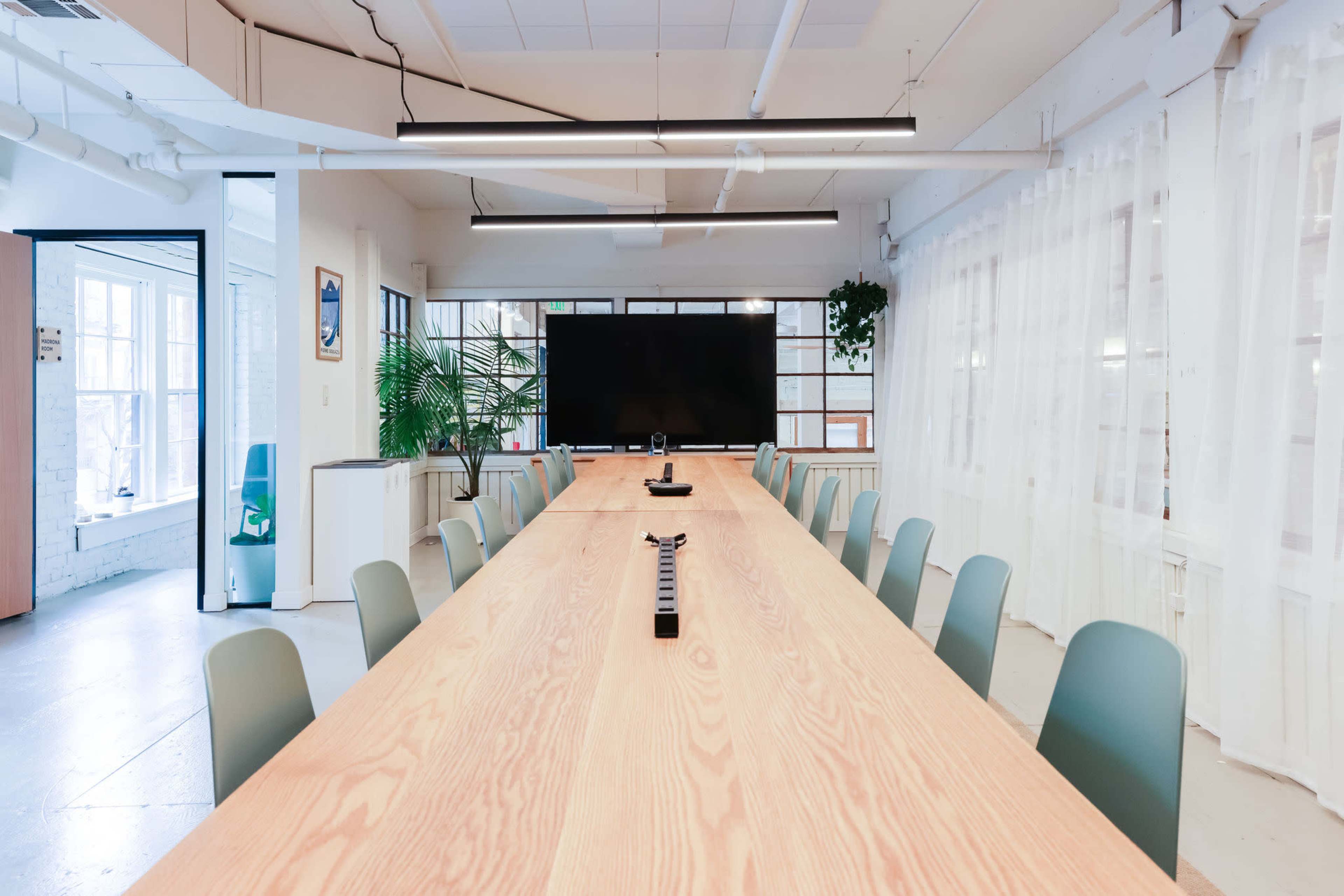 A large, elongated wooden conference table is set up in a bright meeting room with a television screen at one end and chairs lined along both sides.