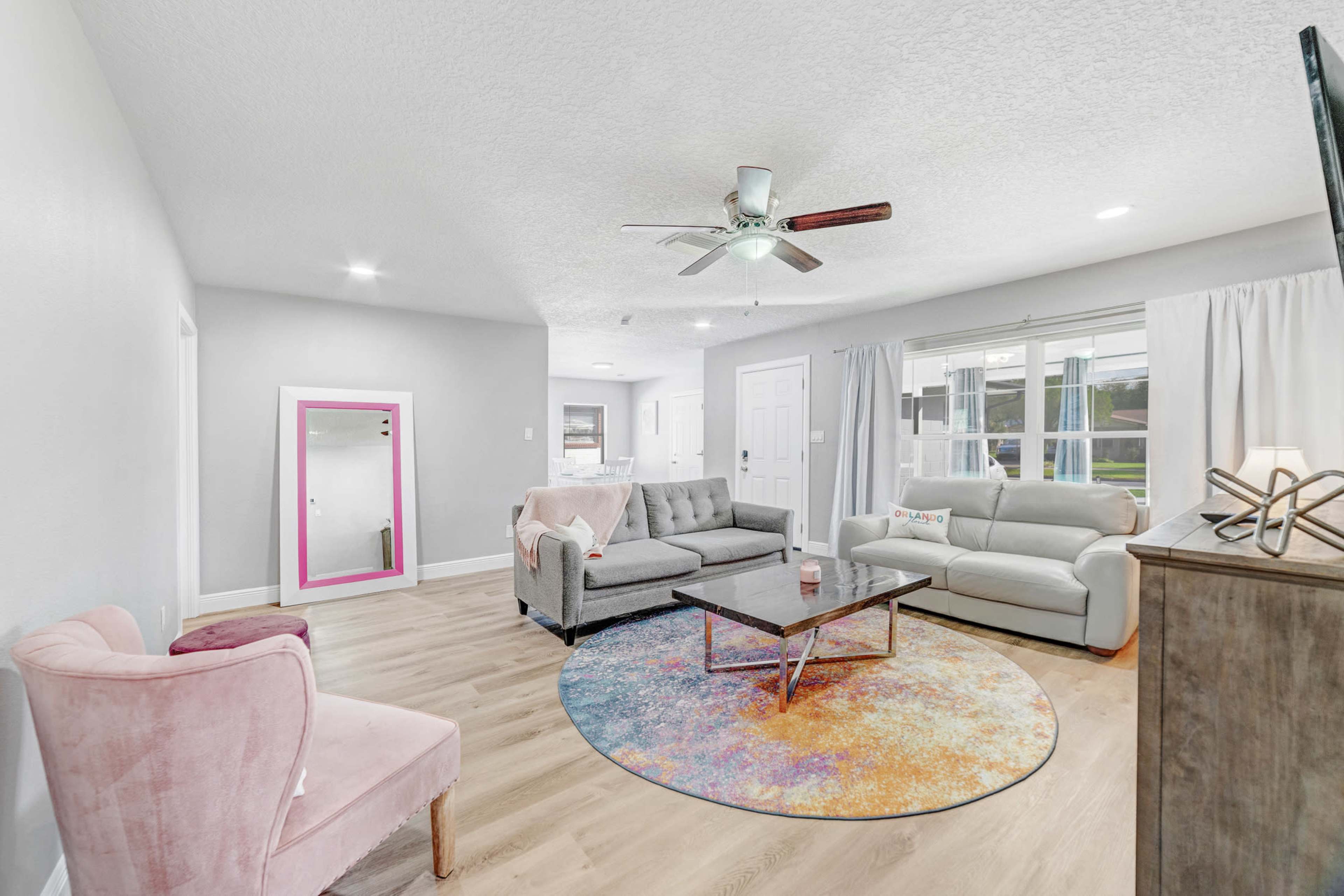 The living room features a light-colored sofa, a gray sectional, a circular rug, and a large mirror with a pink frame against the wall.