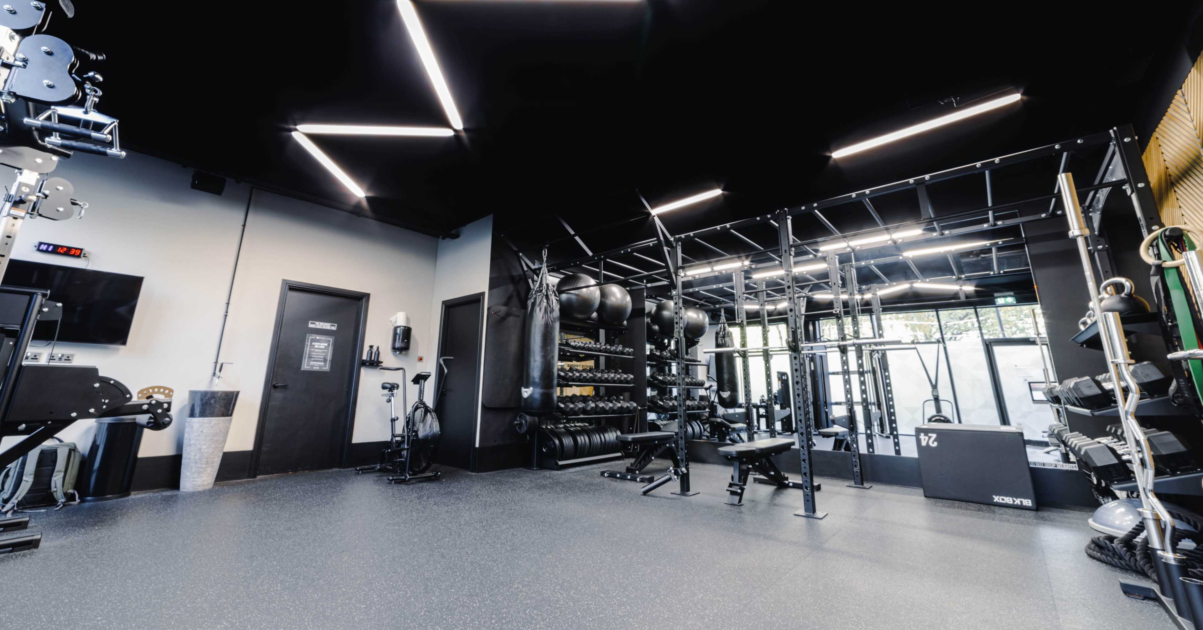 The image shows a modern gym interior featuring various workout equipment, including weight racks, benches, and a functional training rig, set against a sleek black and gray color scheme.