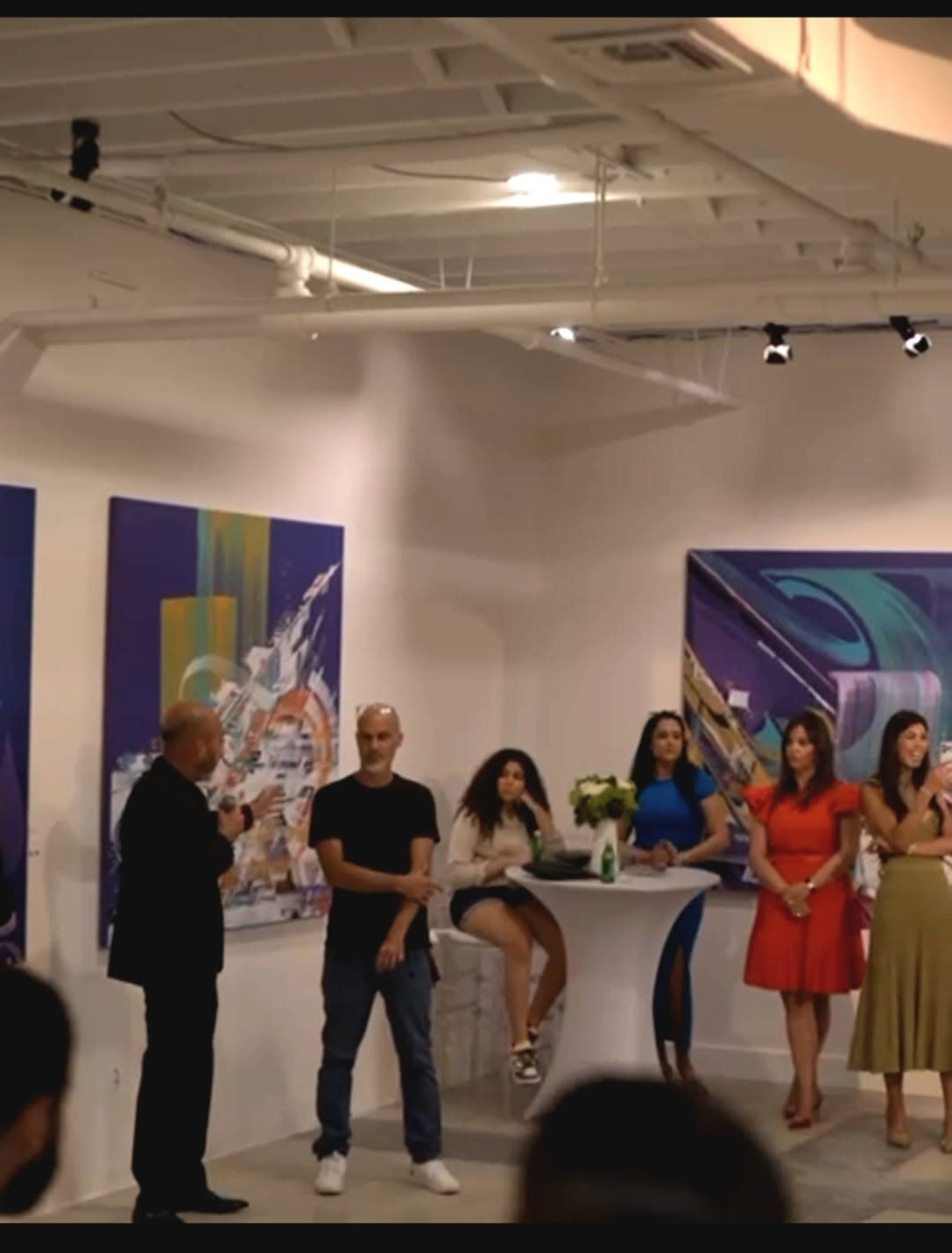A group of people stands in an art gallery, with one man speaking while several others observe and two women sit at a table near colorful abstract paintings on the walls.