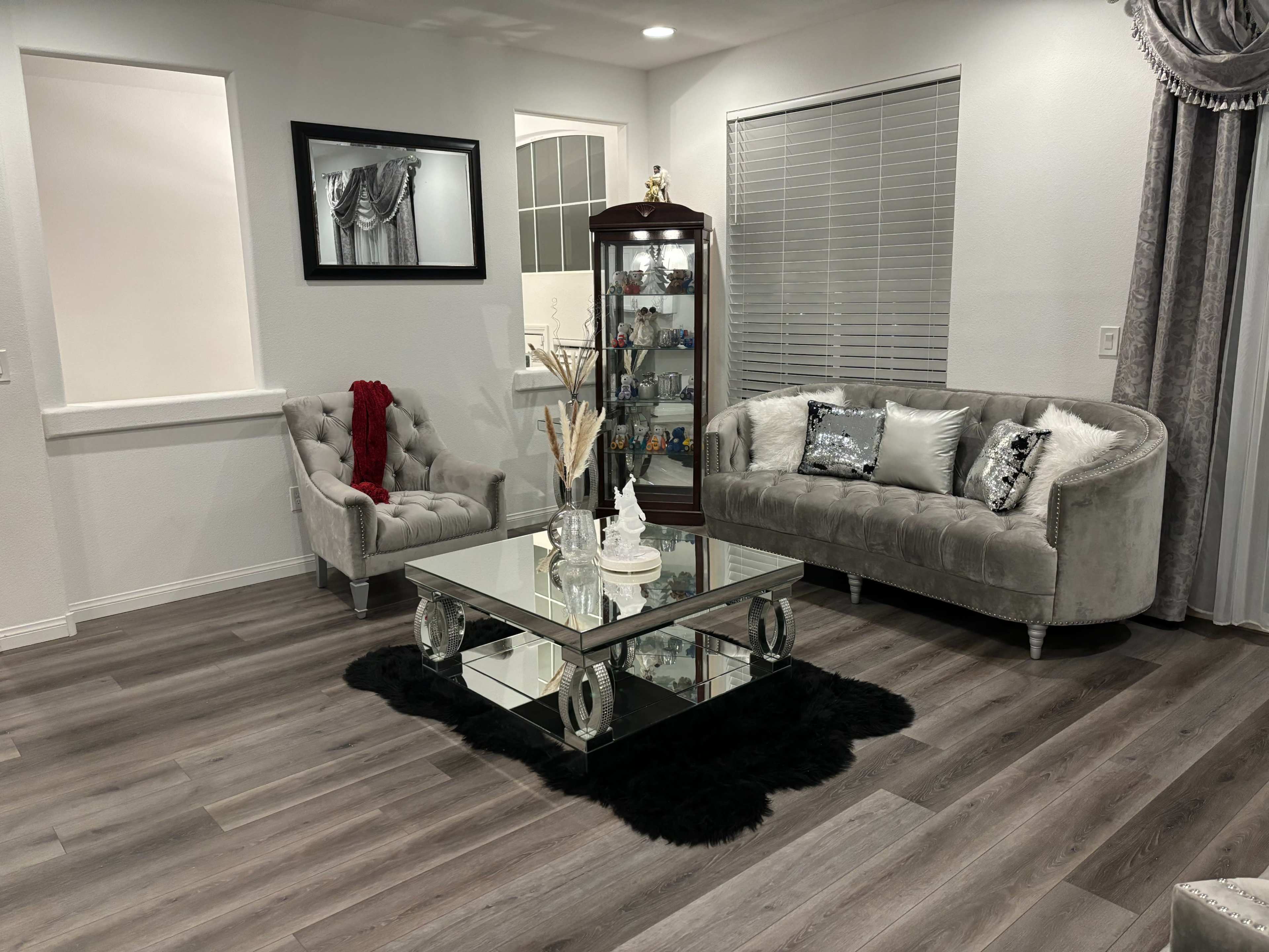The image shows a modern living room featuring a mirrored coffee table, a glass display cabinet, and two gray upholstered sofas arranged on wood flooring.