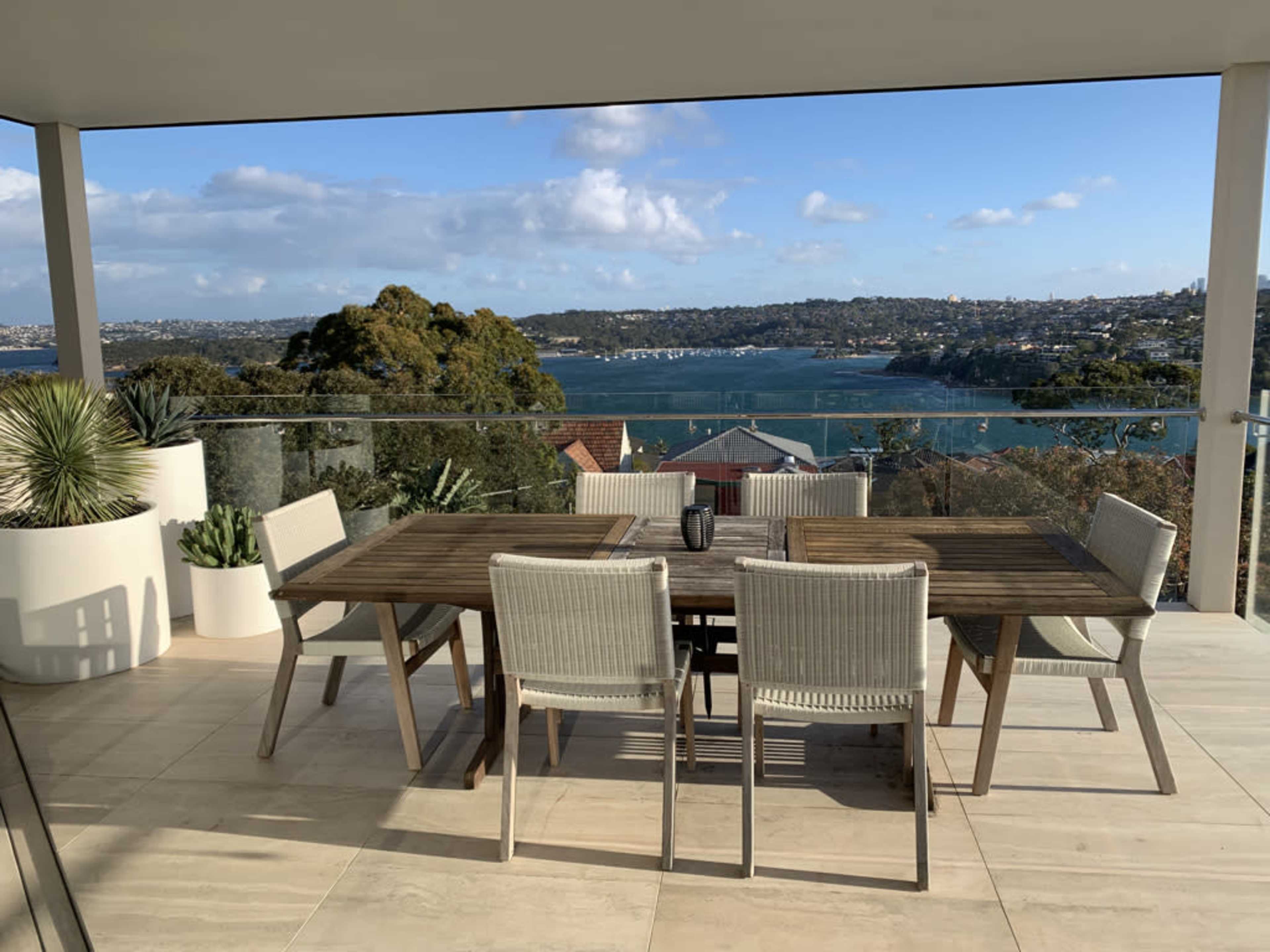 A modern outdoor dining area features a wooden table and six chairs, overlooking a scenic view of a bay and hills.