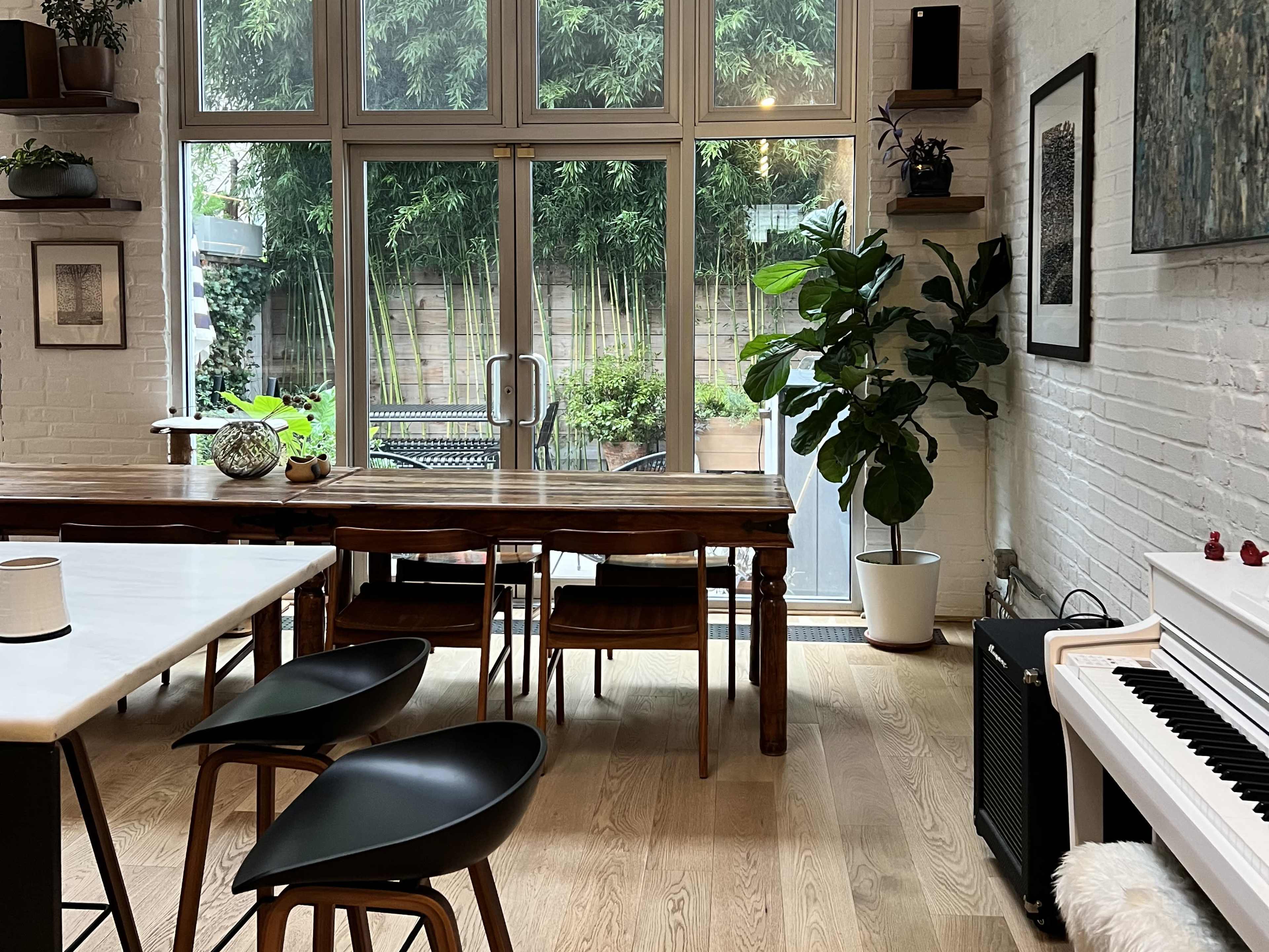 The image shows a modern interior space with a wooden dining table, black chairs, a white piano, and large windows overlooking a green outdoor area.