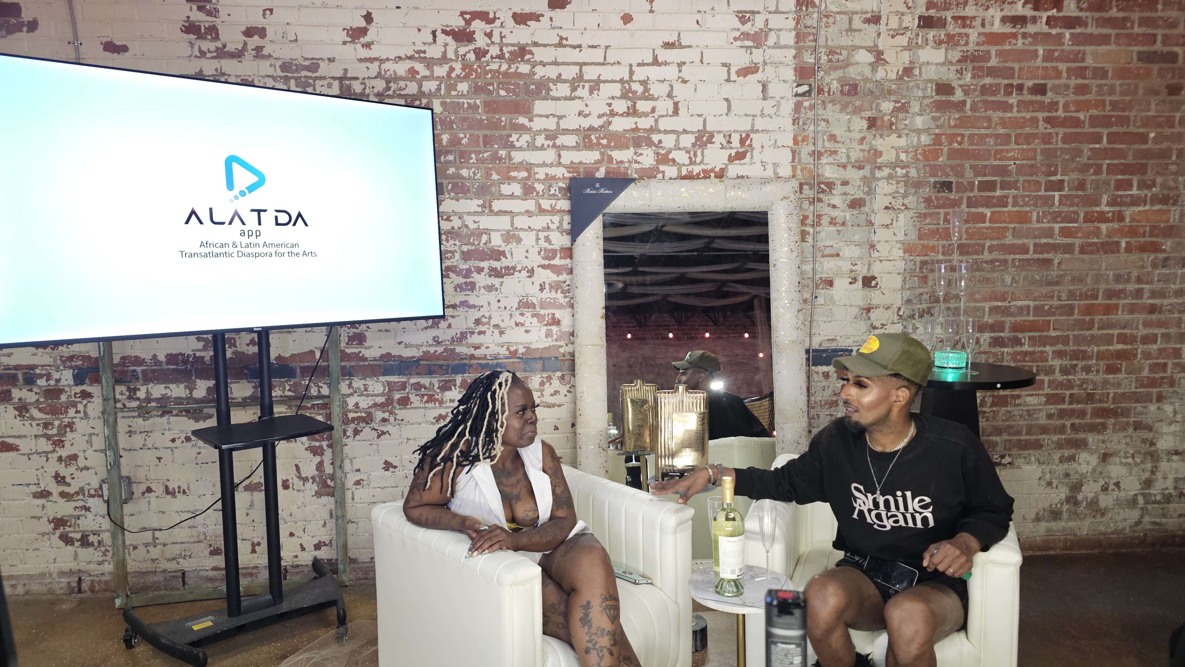 Two individuals are seated in white chairs in front of a large screen displaying the logo for "ALATDA," against a backdrop of exposed brick walls.