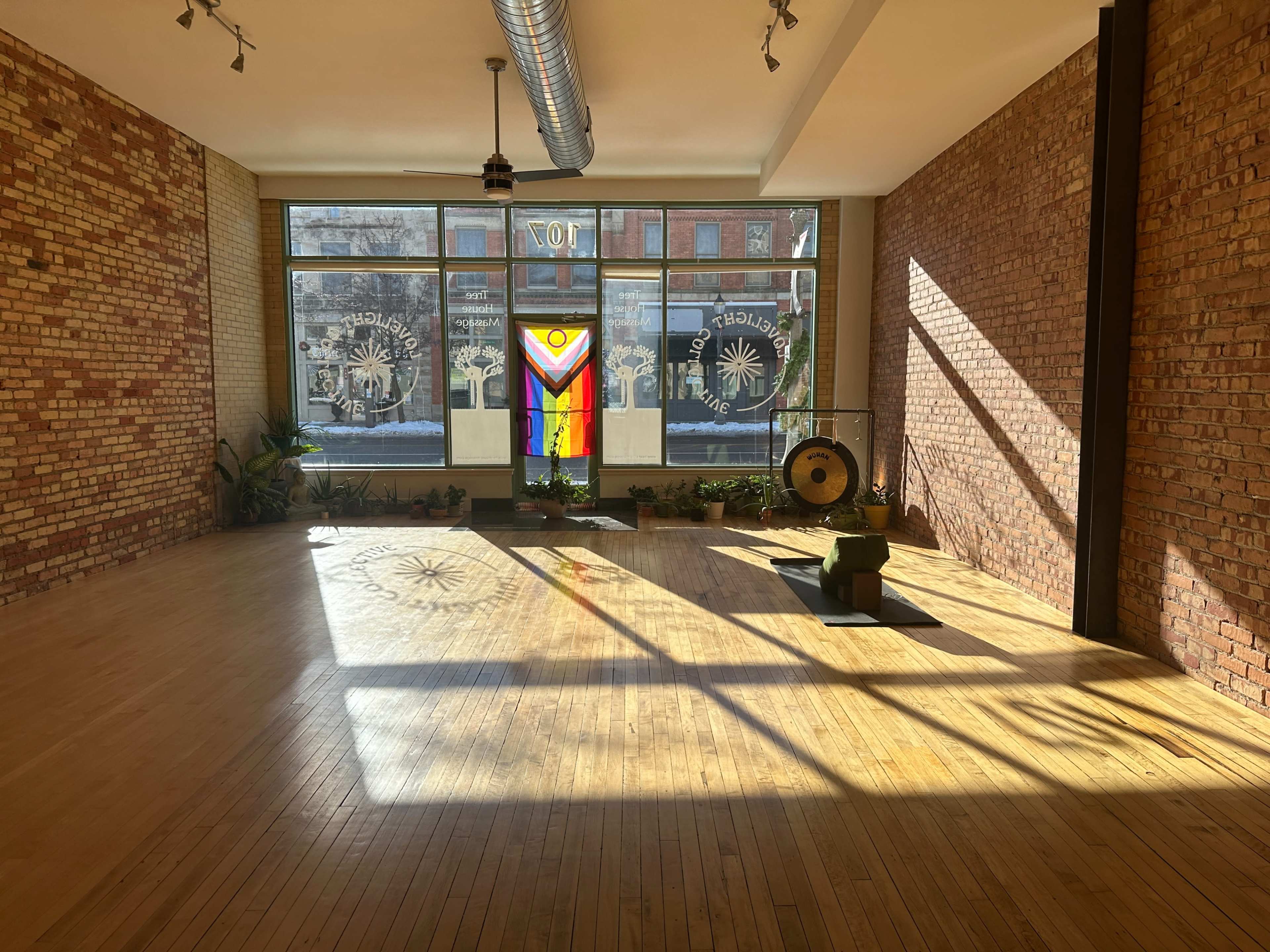 The image shows a spacious, sunlit room with wooden flooring, large windows featuring a rainbow flag, and a gong positioned in a corner.
