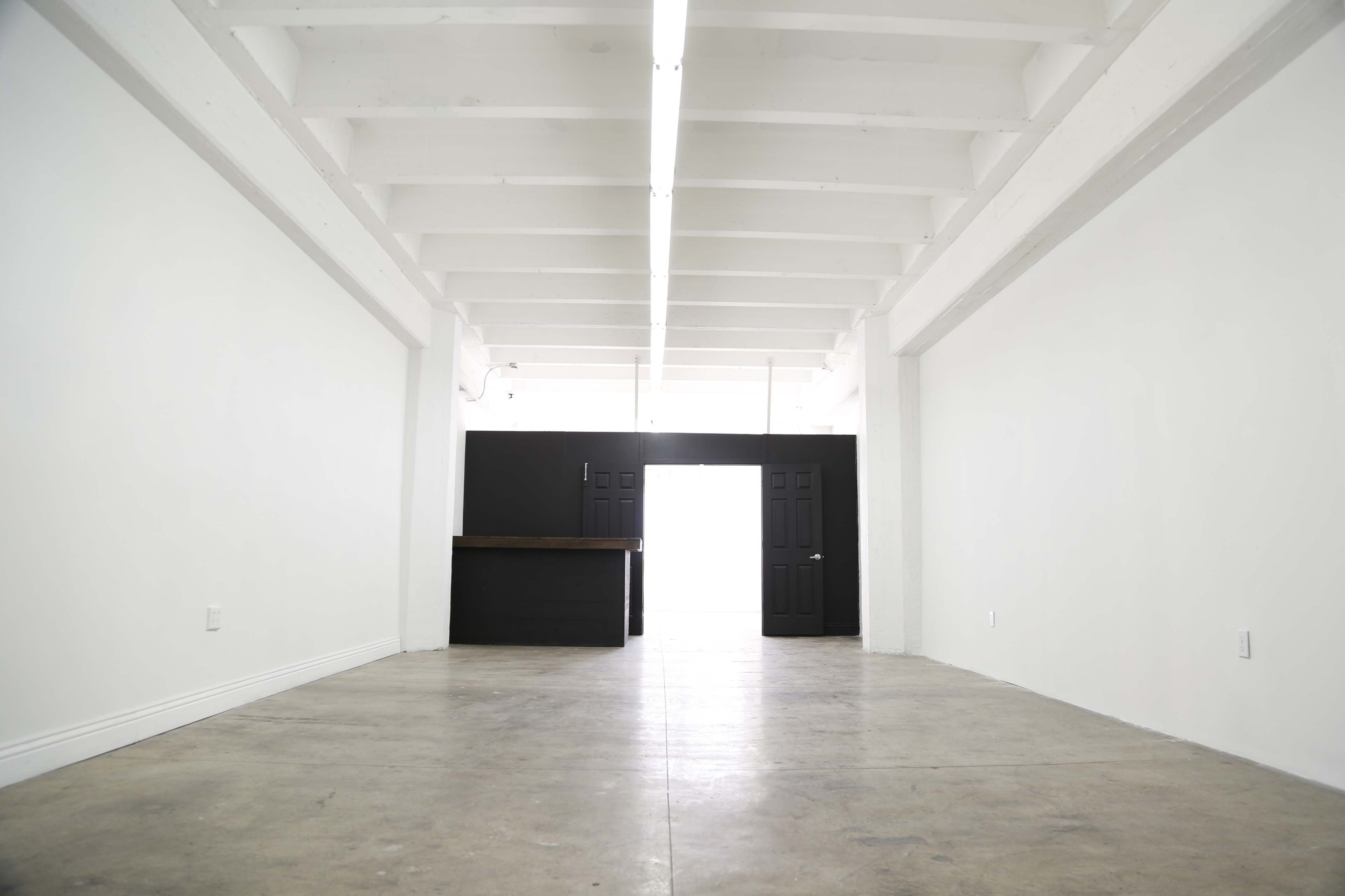 The image shows an empty room with white walls, a concrete floor, and a small black counter near the back.