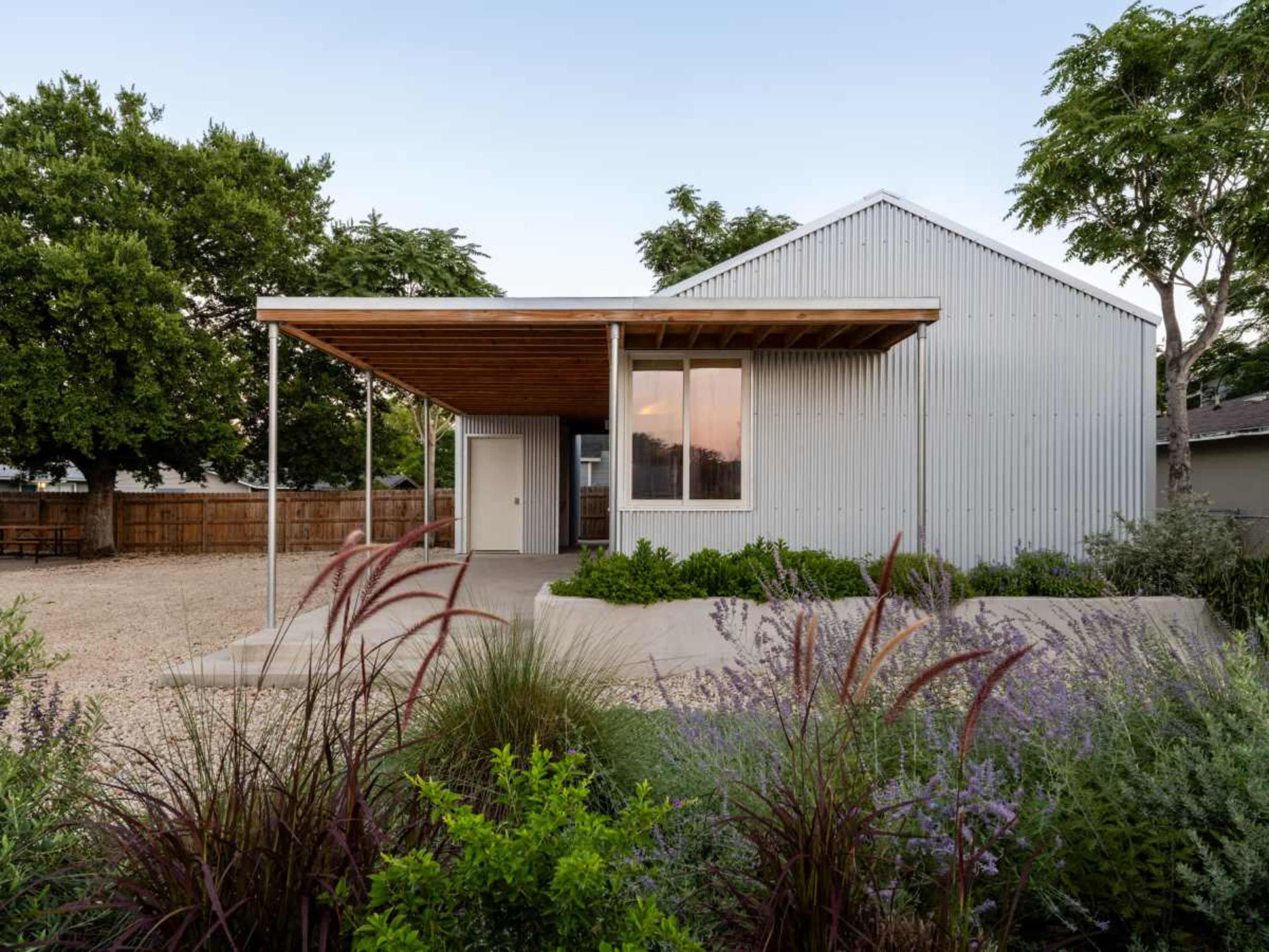 A modern house with a metal exterior and wooden overhang is surrounded by landscaping featuring various plants and a gravel driveway.