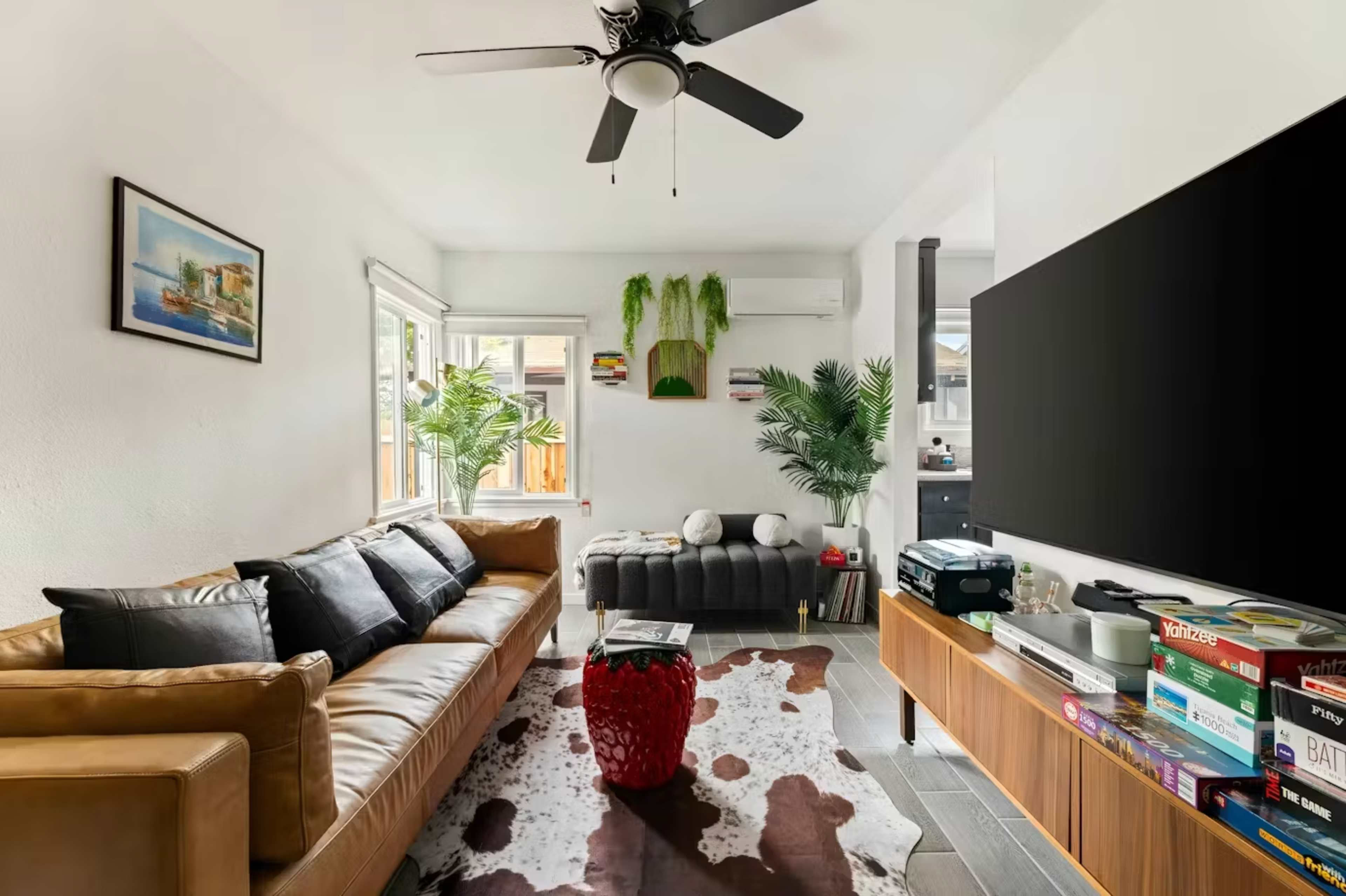 A modern living room features a leather sofa, a black ottoman, a large TV on a wooden stand, and potted plants near the windows.