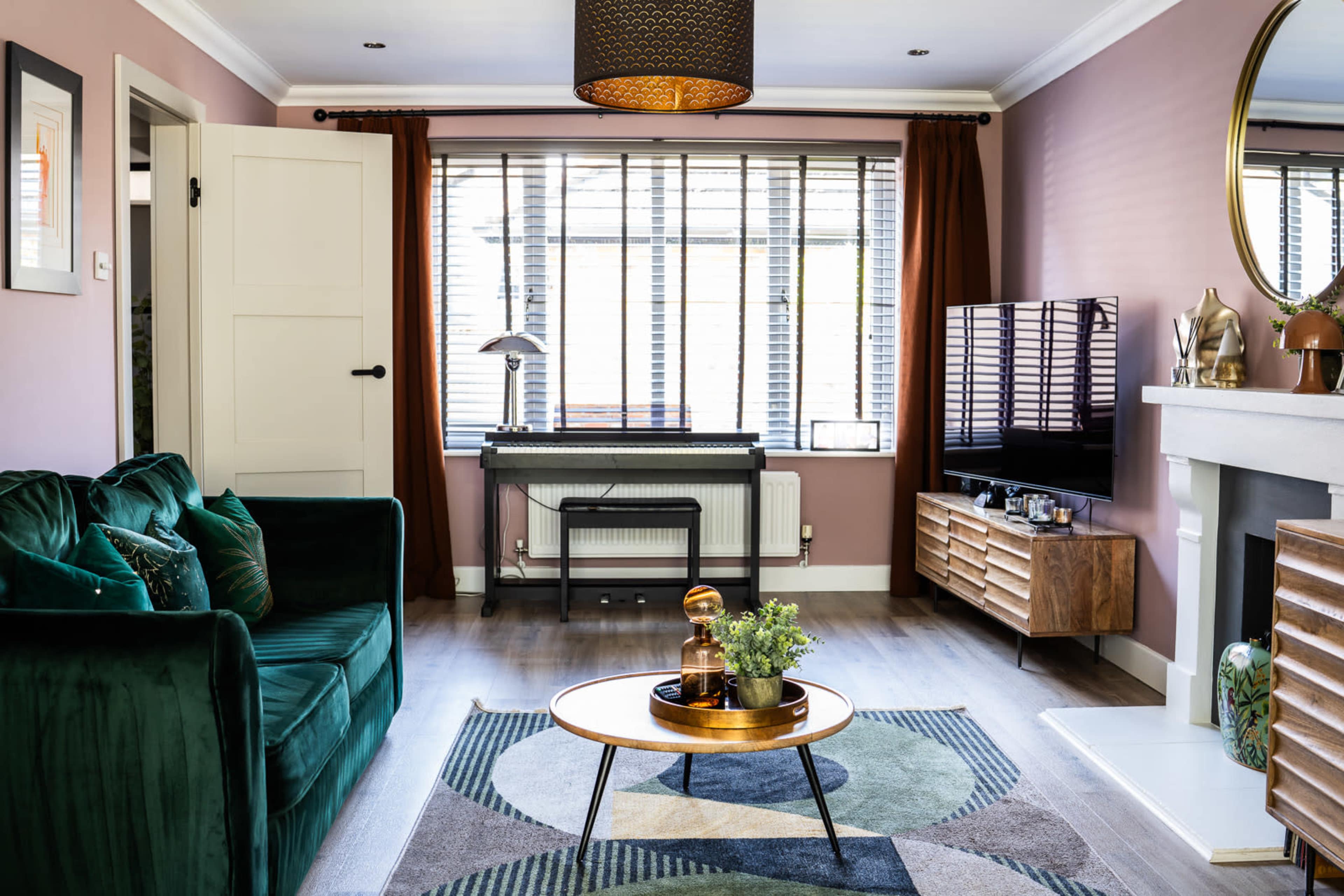 The image shows a modern living room featuring a green velvet sofa, a round coffee table, a TV stand, and large windows with blinds.