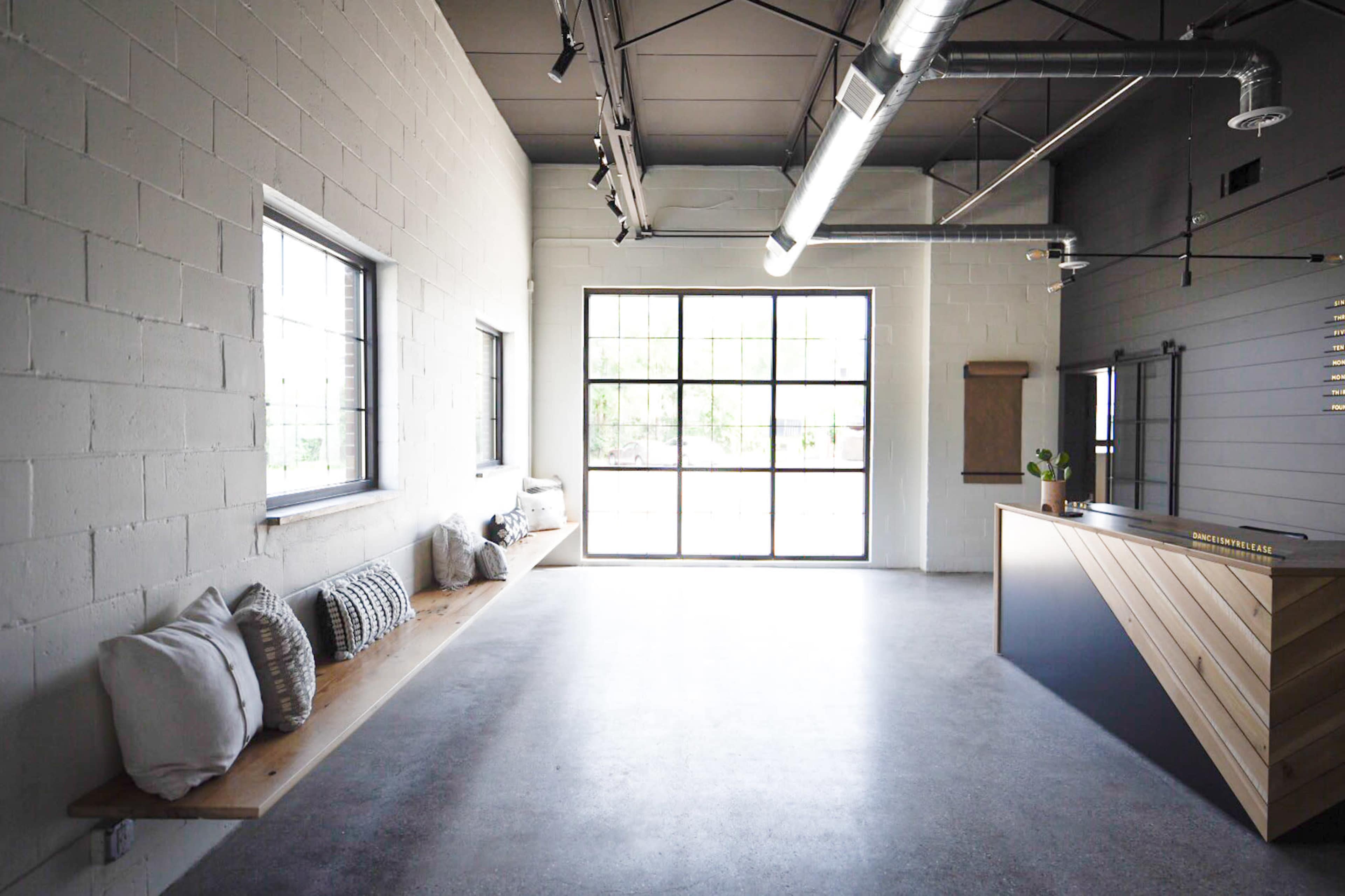 A minimalist reception area featuring a long bench with cushions, large windows, and a wooden front desk.