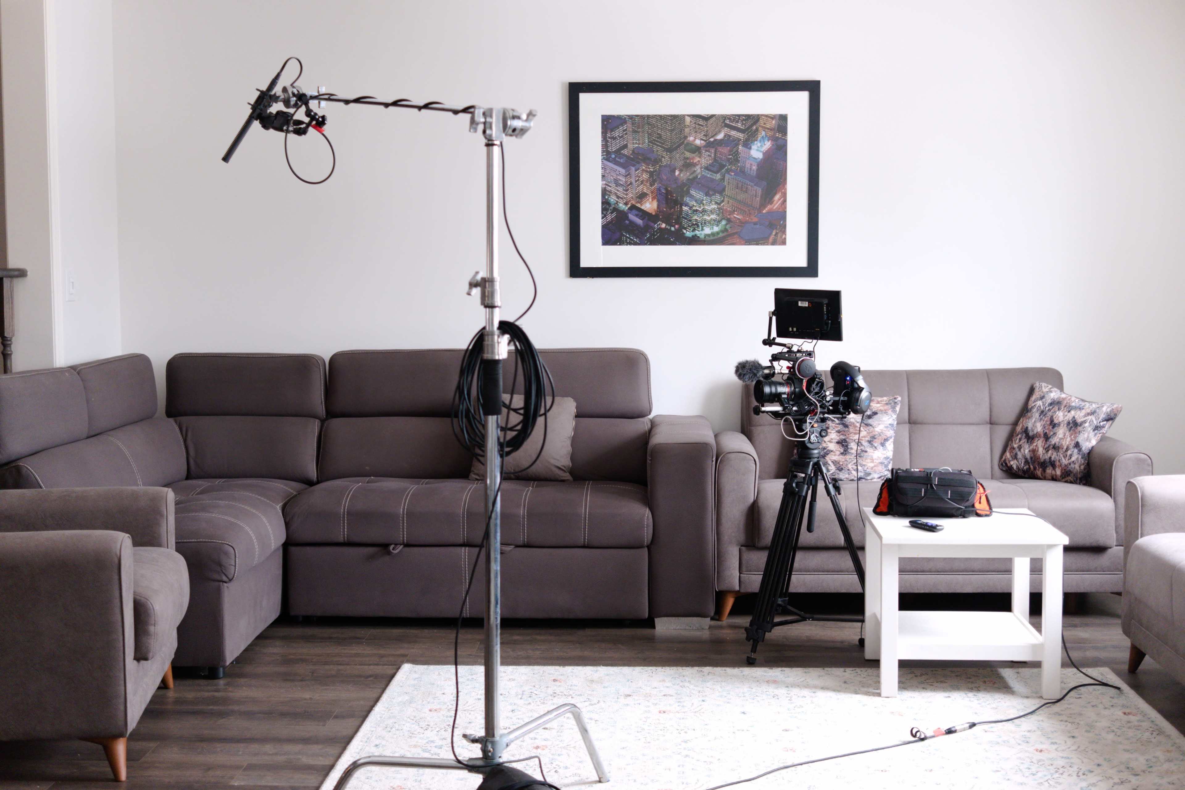 A living room is set up for a film shoot with a camera on a tripod, a microphone on a boom stand, and two couches arranged around a coffee table.