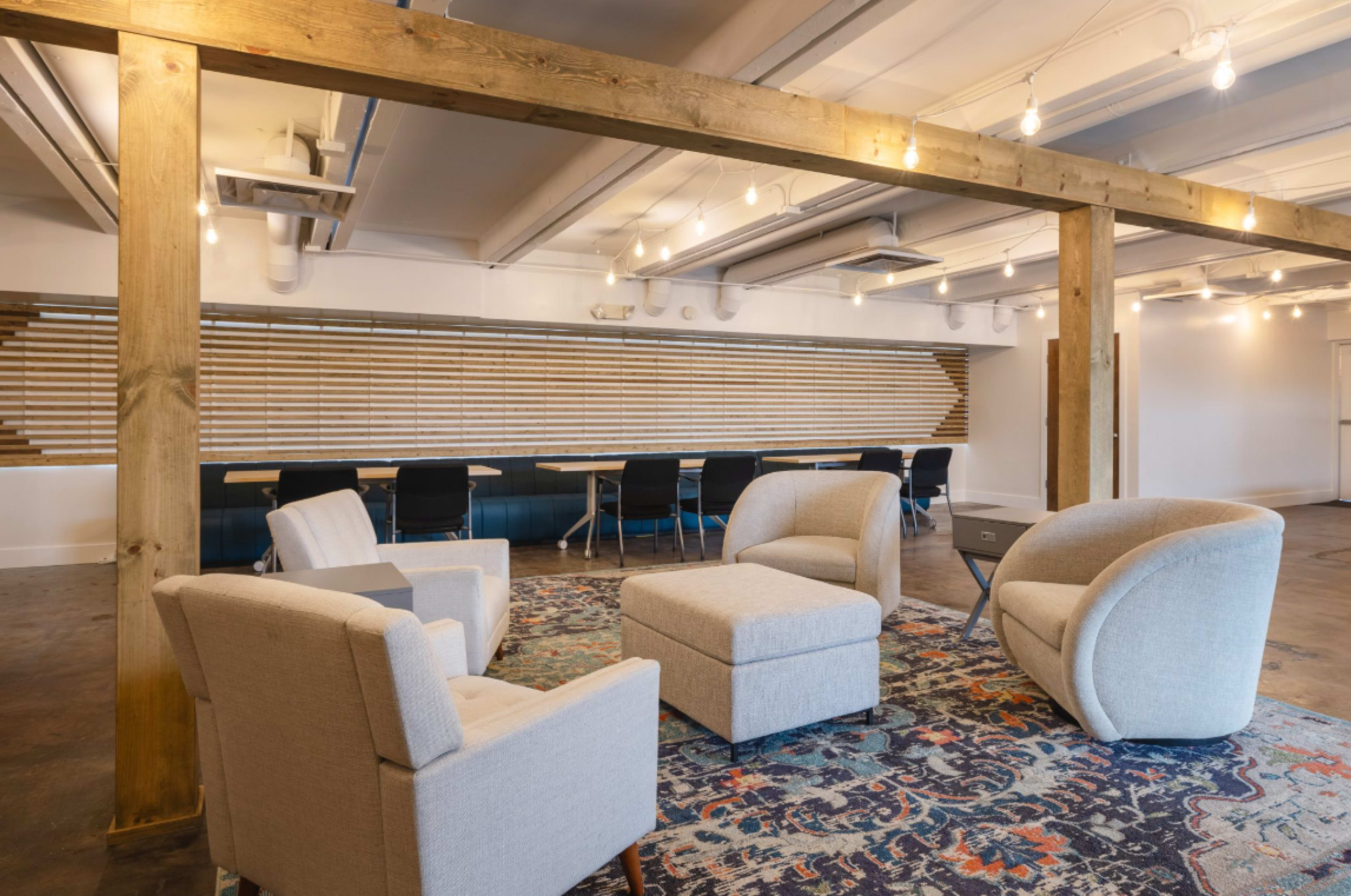 The image shows a modern office space with a seating area featuring two armchairs and an ottoman on a patterned rug, alongside a long table and black chairs in the background.
