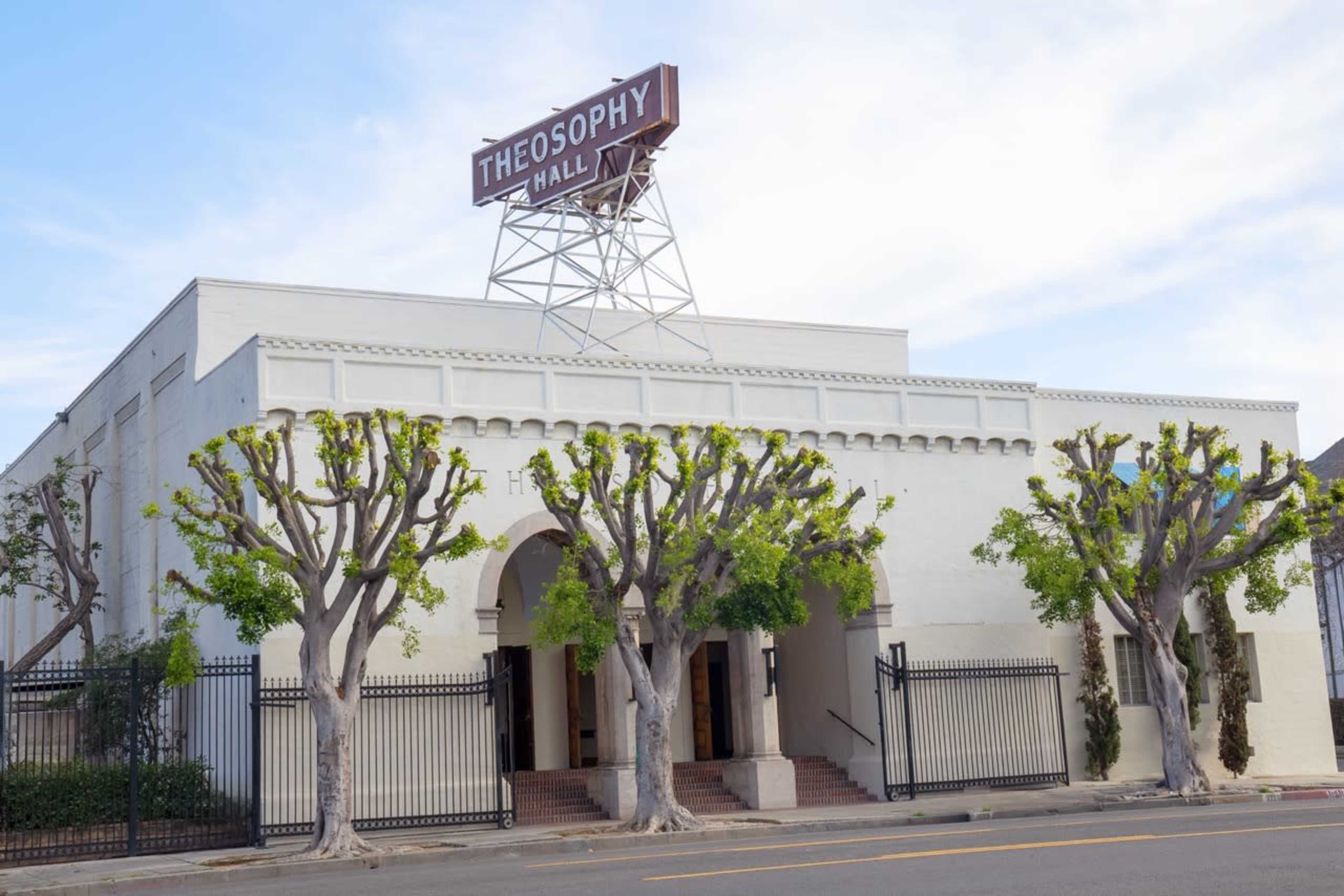 The image shows Theosophy Hall, a large white building with a prominent sign, framed by green trees and a gate.