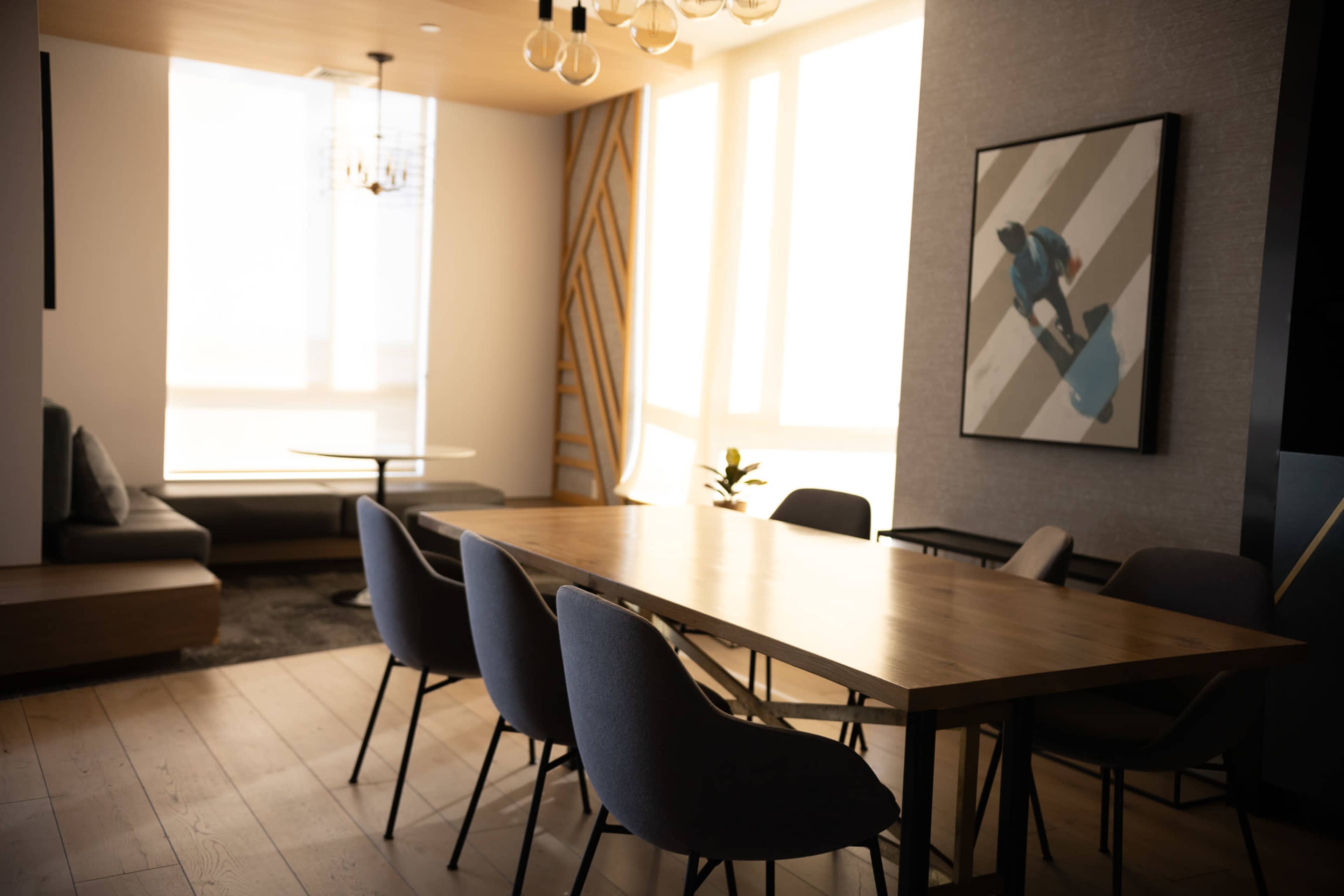 A modern meeting room features a large wooden table surrounded by upholstered chairs, with a wall art piece and sunlight streaming through large windows.