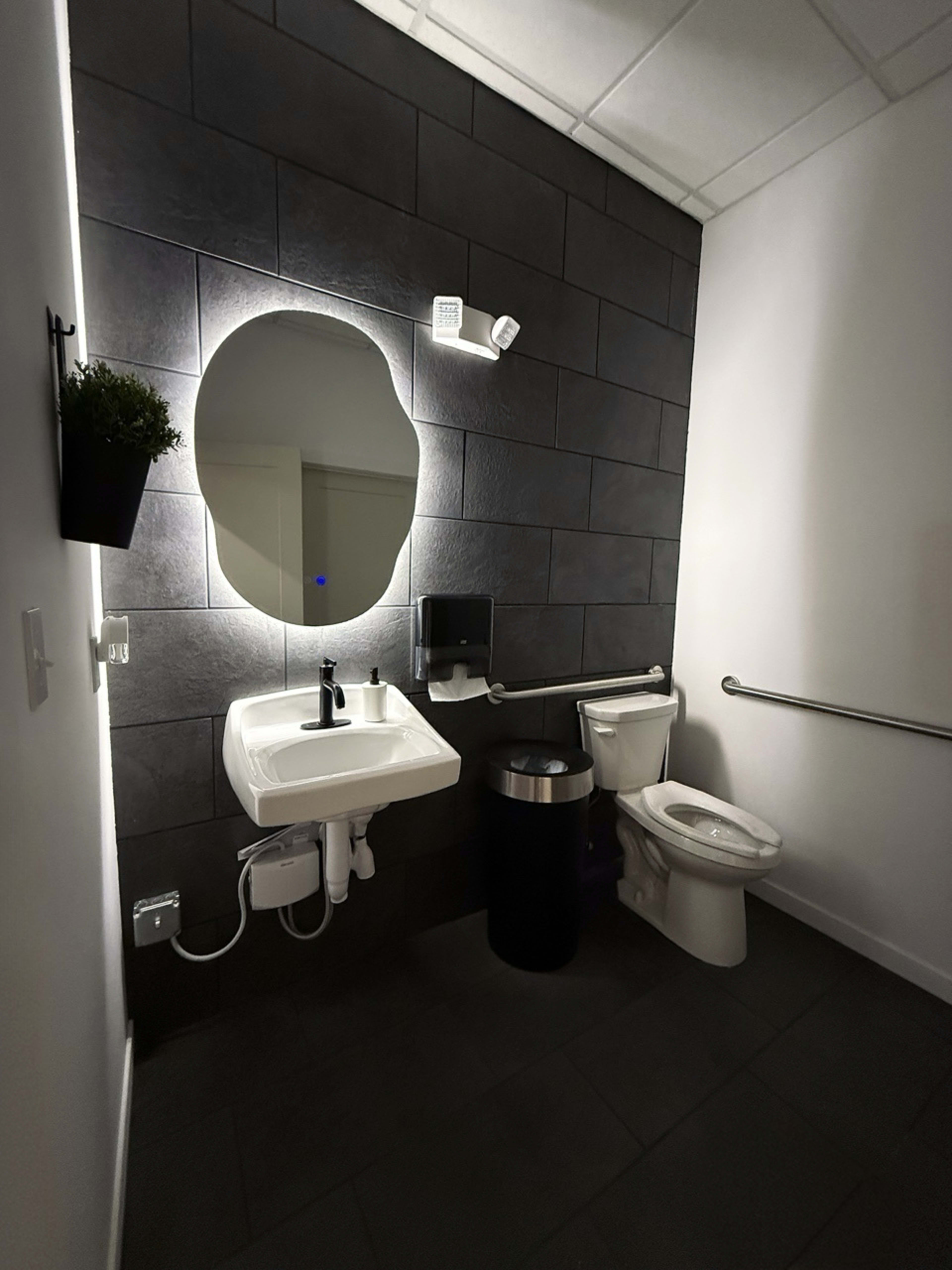 The image shows a modern restroom with a wall-mounted sink, a round mirror, a toilet, and a wall-mounted hand dryer against a dark tile background.