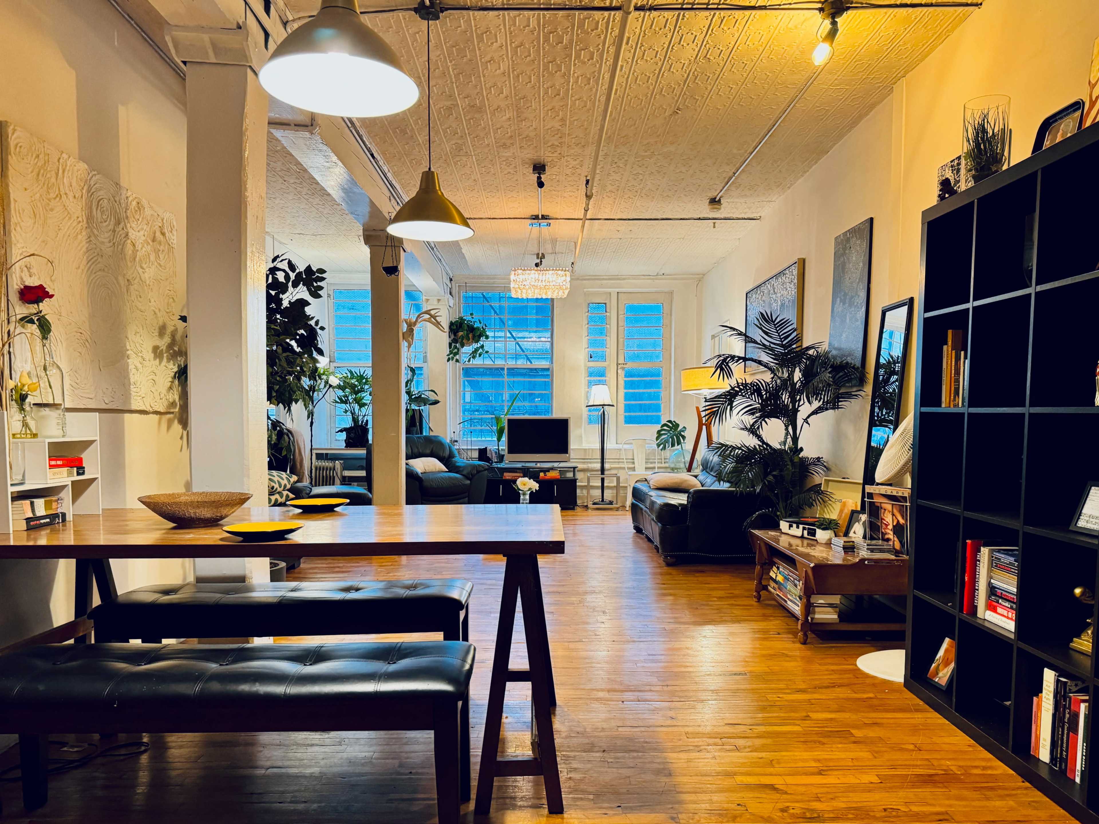 A spacious modern living area features a dining table, black couches, plants, and large windows allowing natural light.