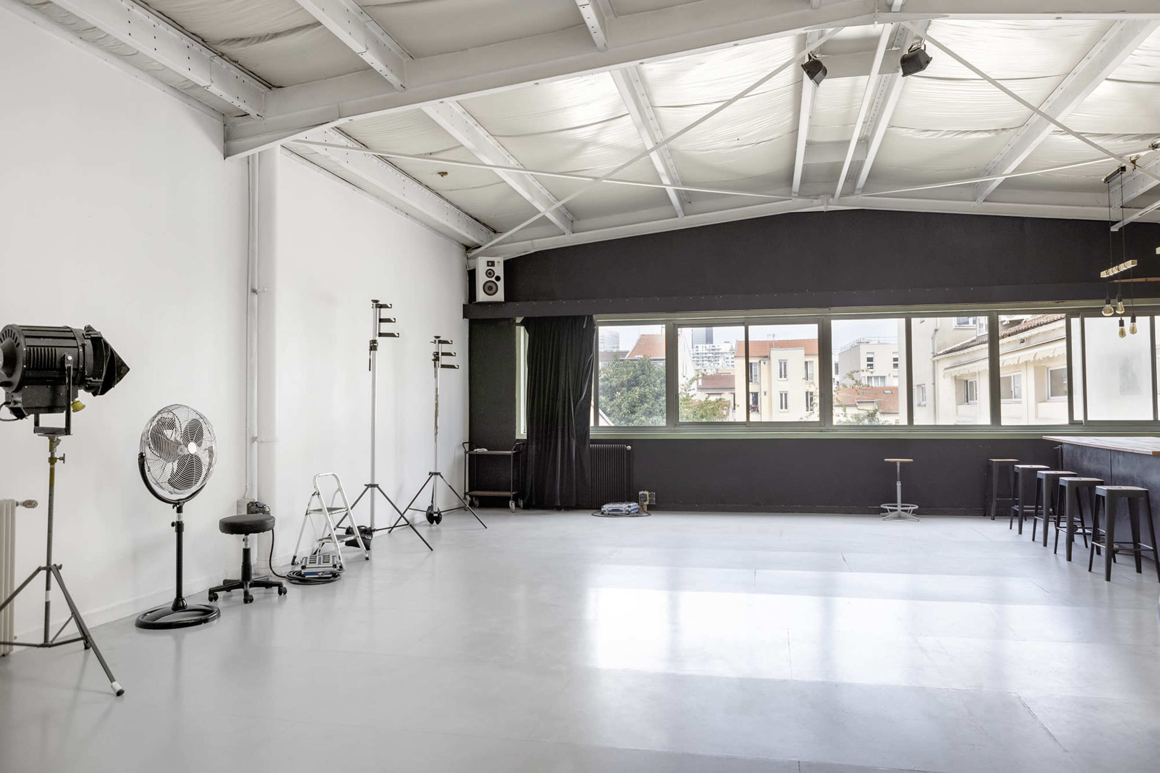 The image shows a spacious, minimally furnished photography studio with large windows and various equipment arranged along the walls.