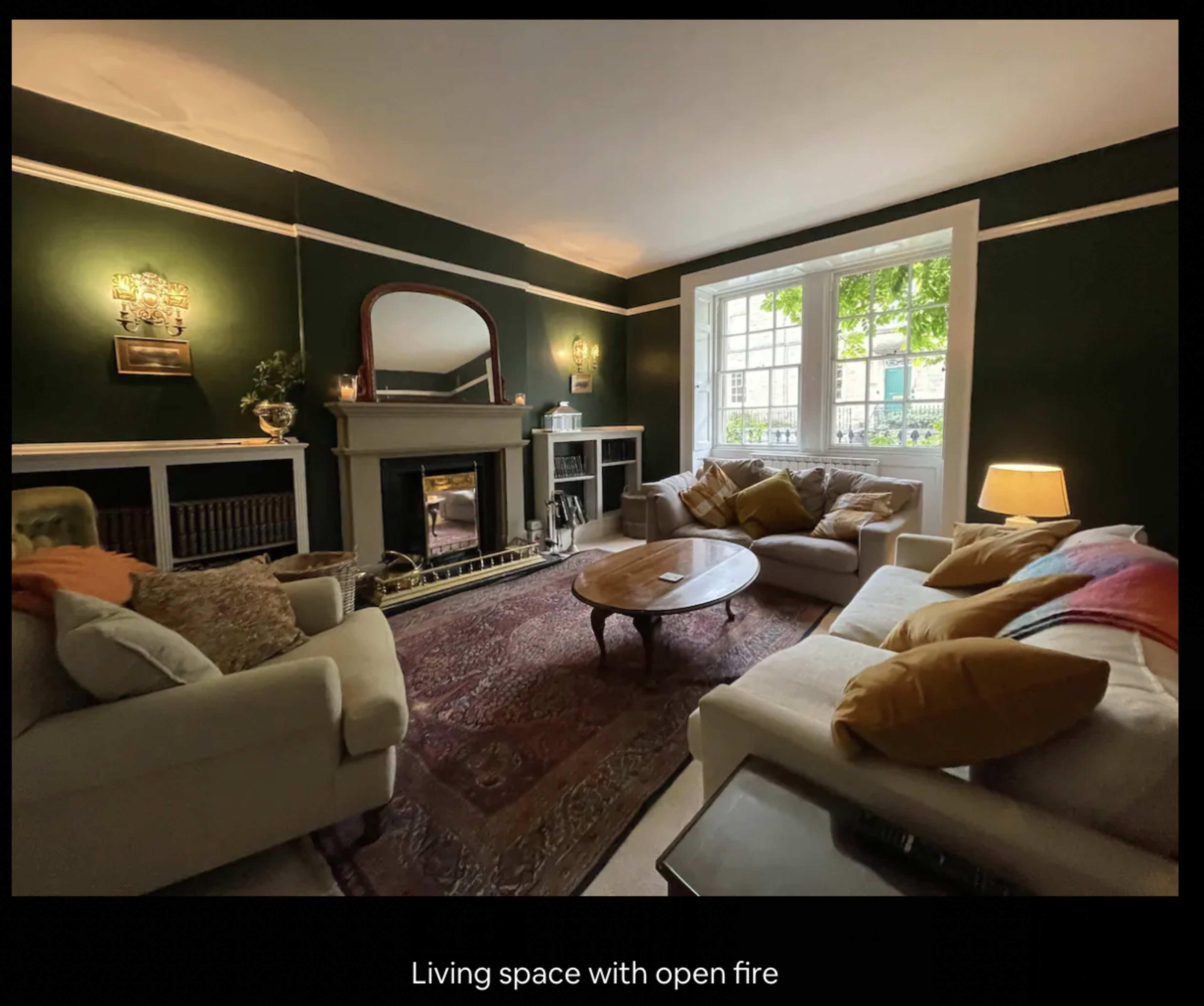 A living room with dark green walls, a large window, a fireplace, and two couches arranged around a coffee table.