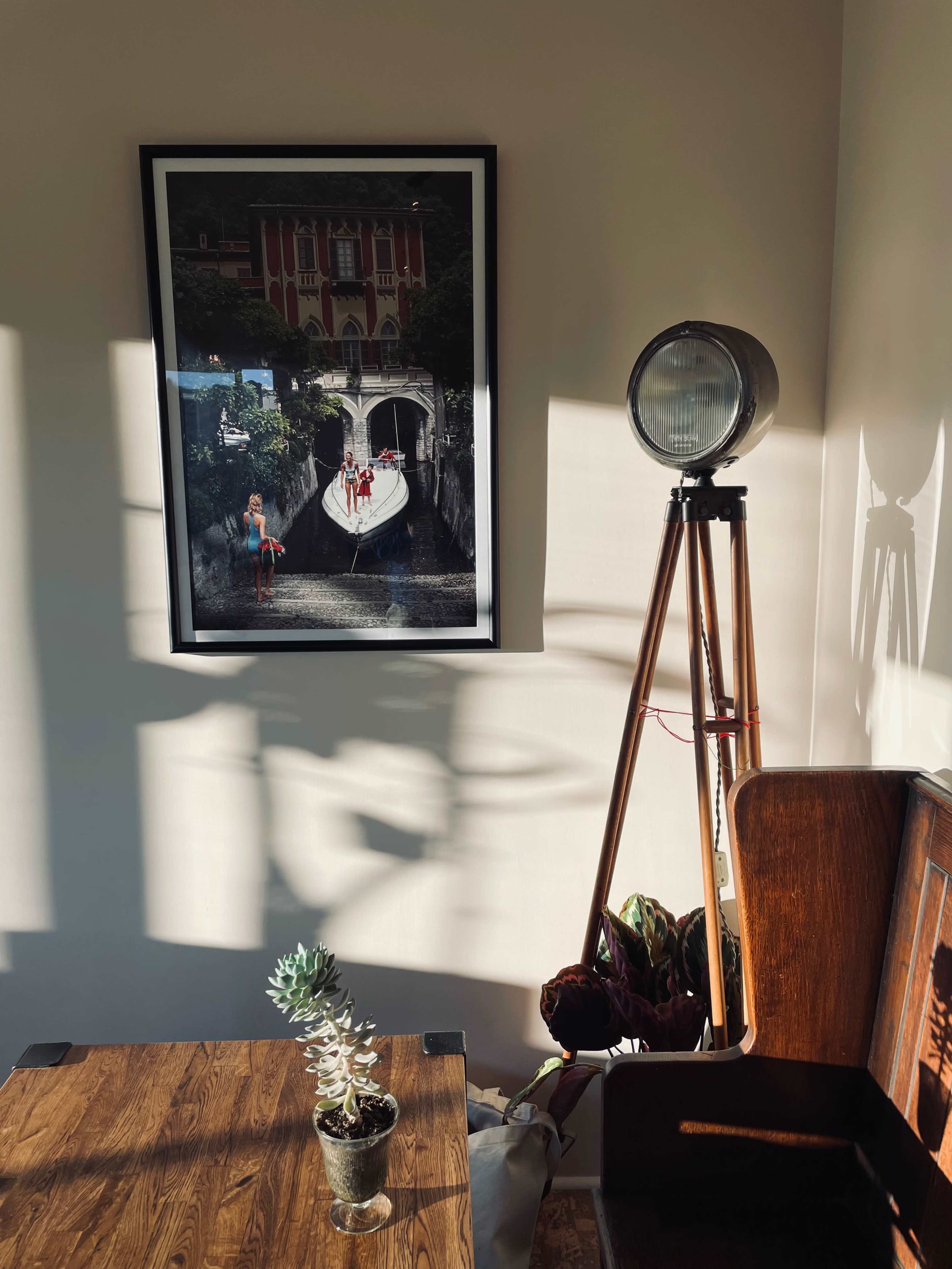 A framed photograph of a boat with people rests on a wall, beside a tripod lamp and a small succulent plant on a wooden table.