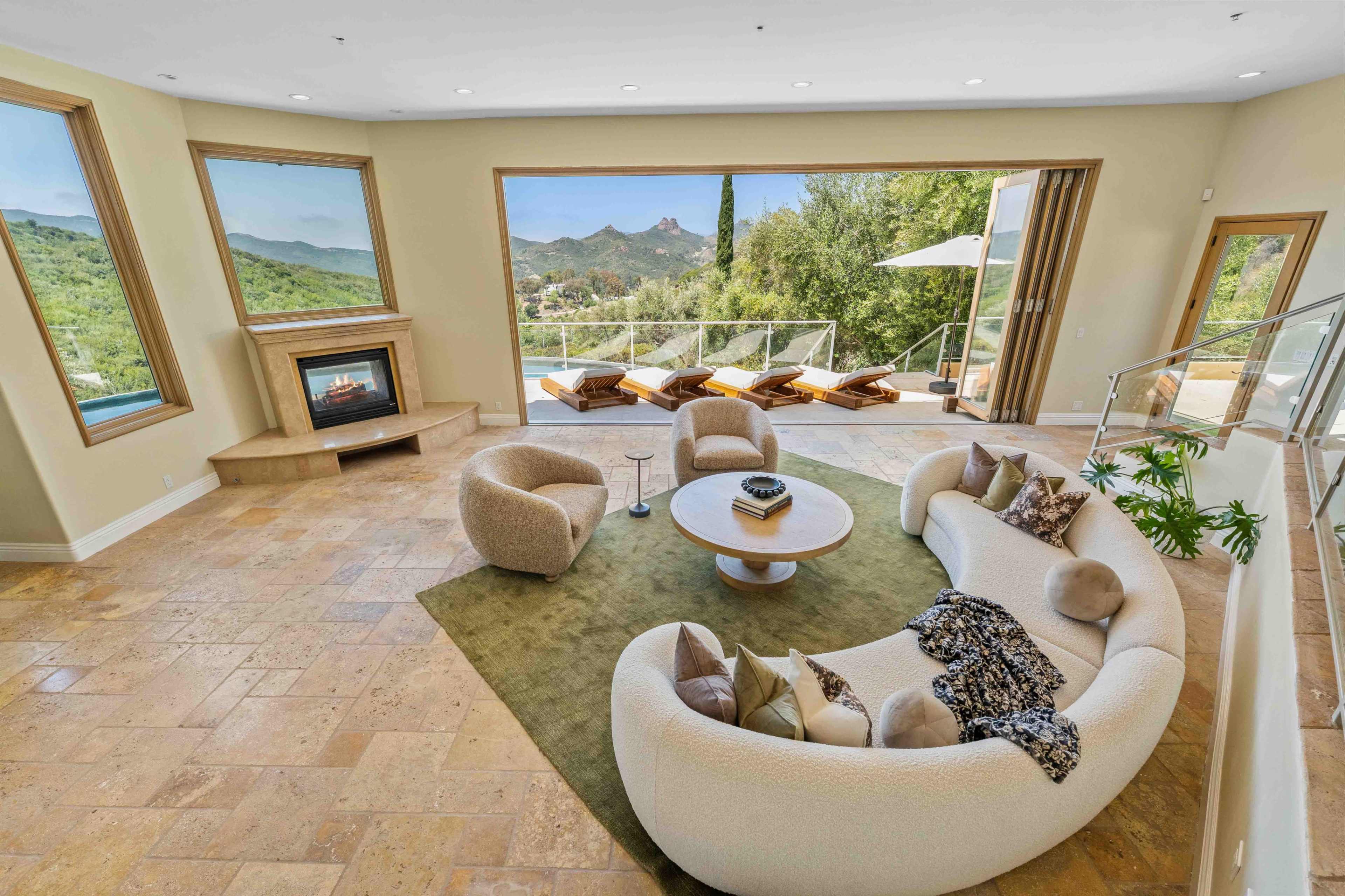 The image shows a modern living room featuring a round white sofa, a circular table, a fireplace, and large windows with views of mountains and greenery.
