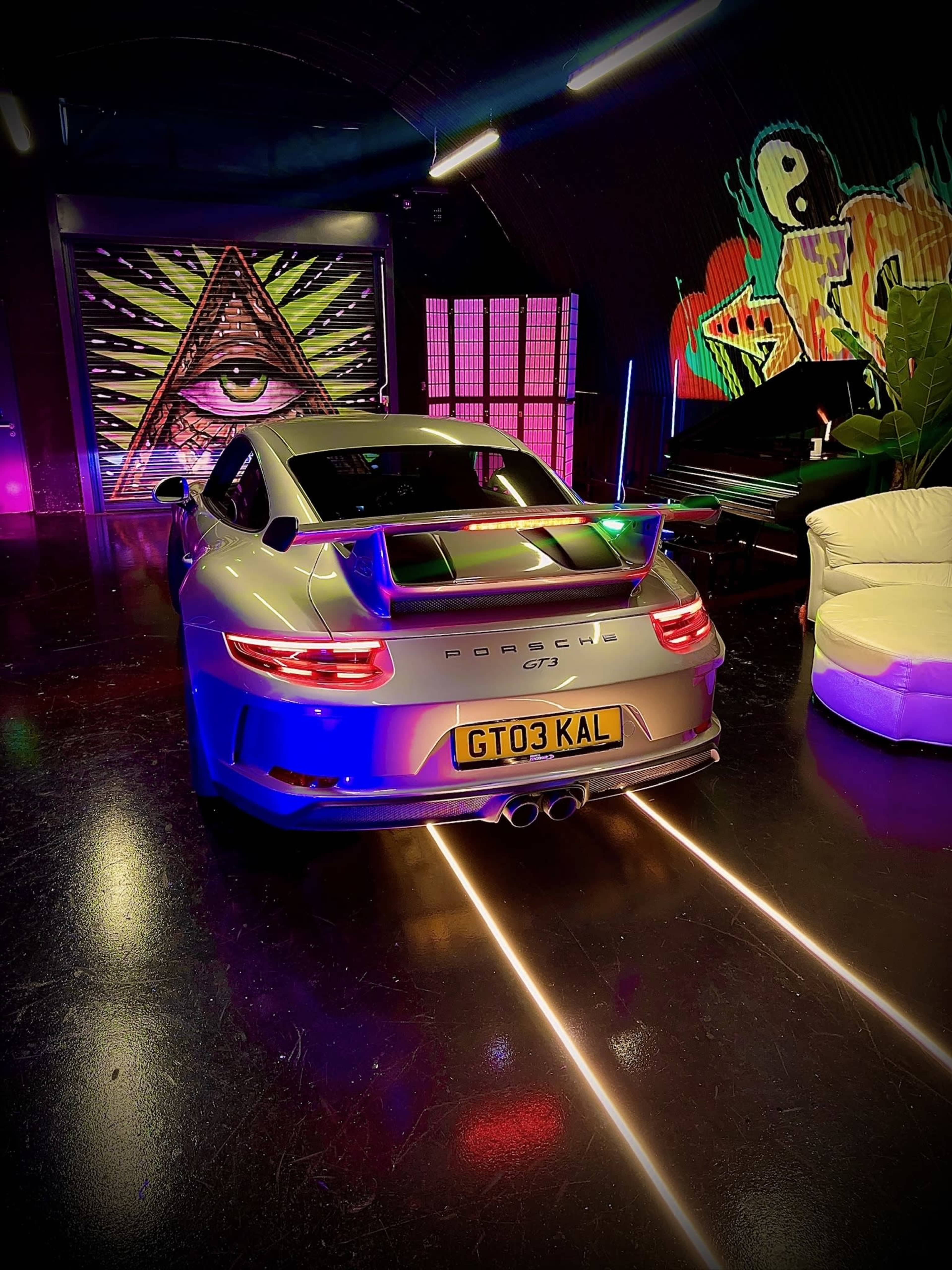 A silver Porsche GT3 appears in a dimly lit room with colorful neon lights and a vibrant mural on the wall.