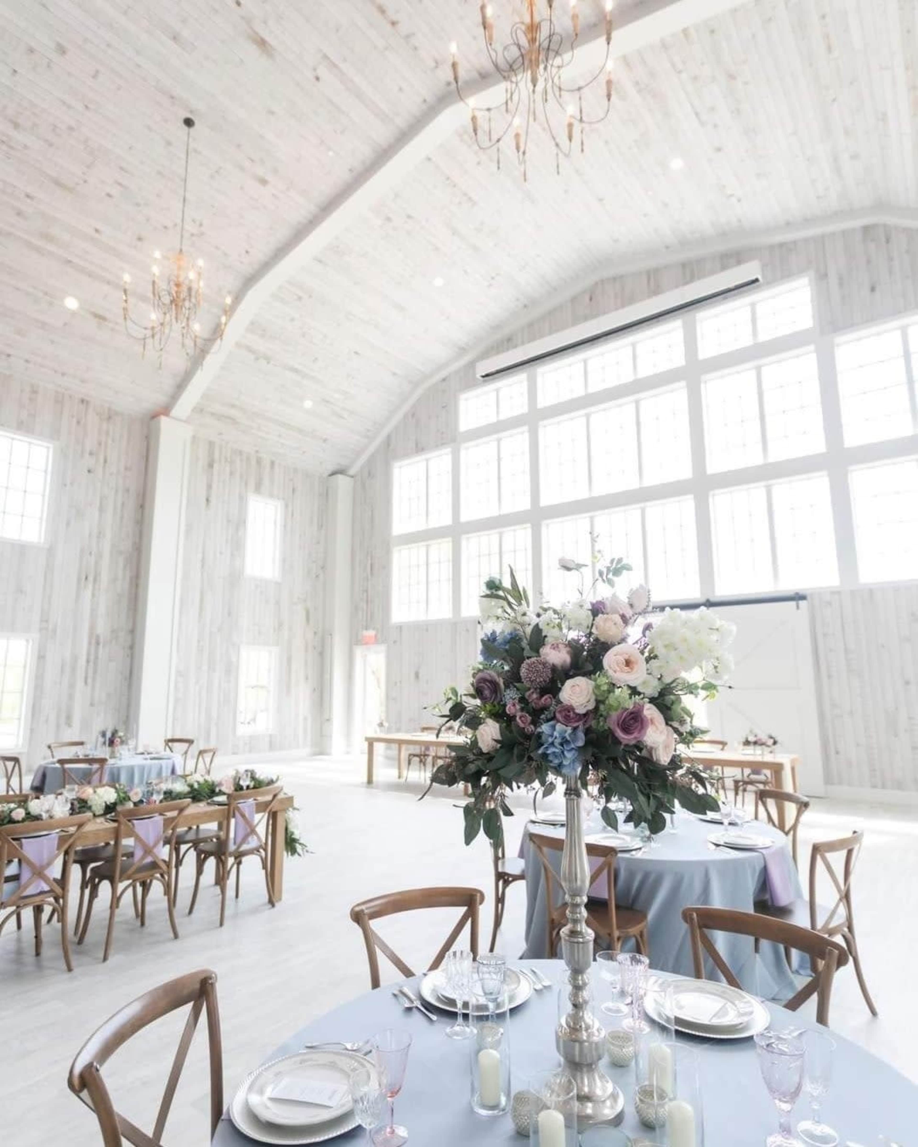 A large, elegantly decorated event space features tall windows, wooden walls, and a floral centerpiece on a table set for dining.