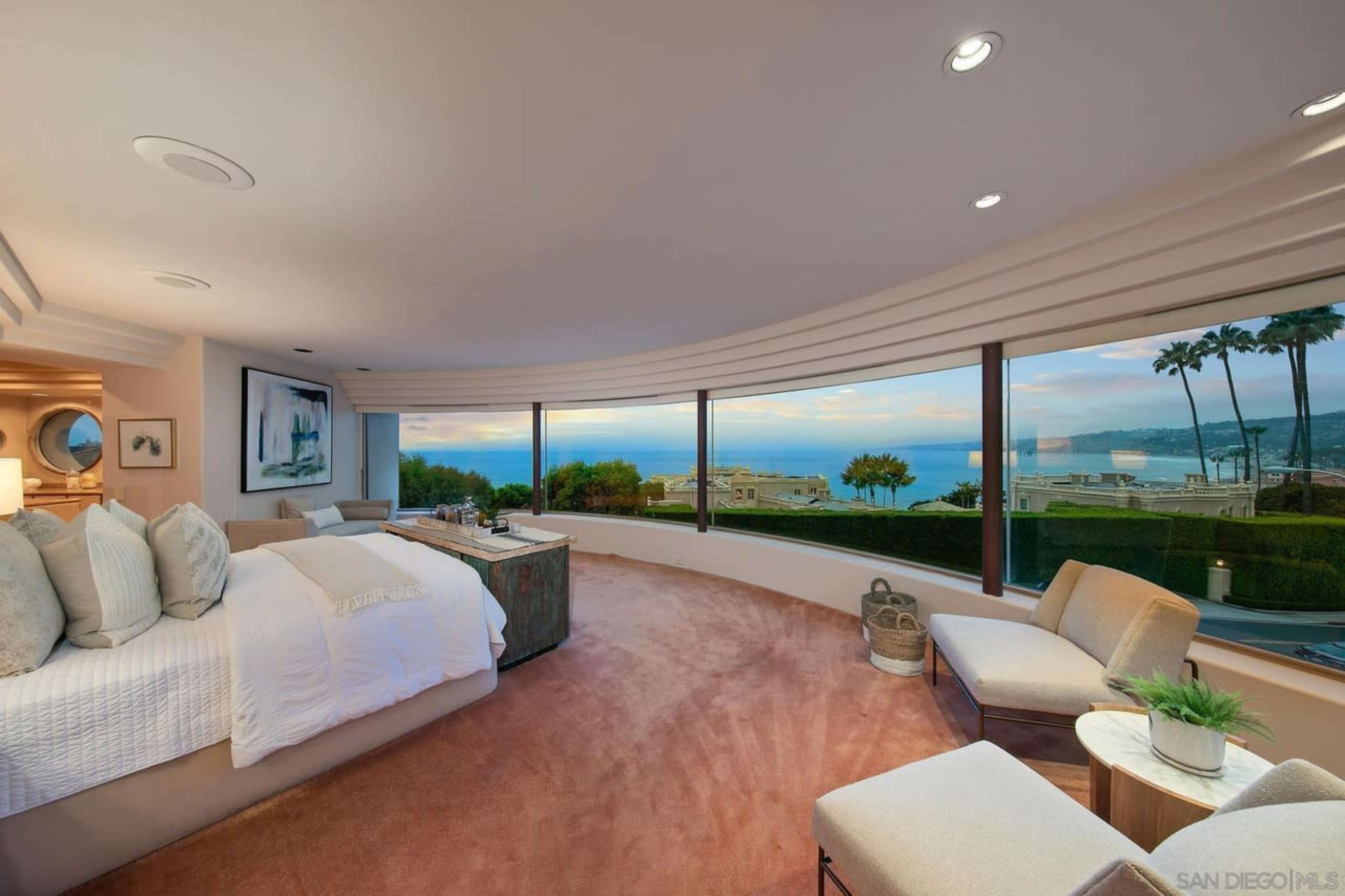 The room features a curved wall of windows overlooking a coastal view, with a large bed, a seating area, and decorative elements.
