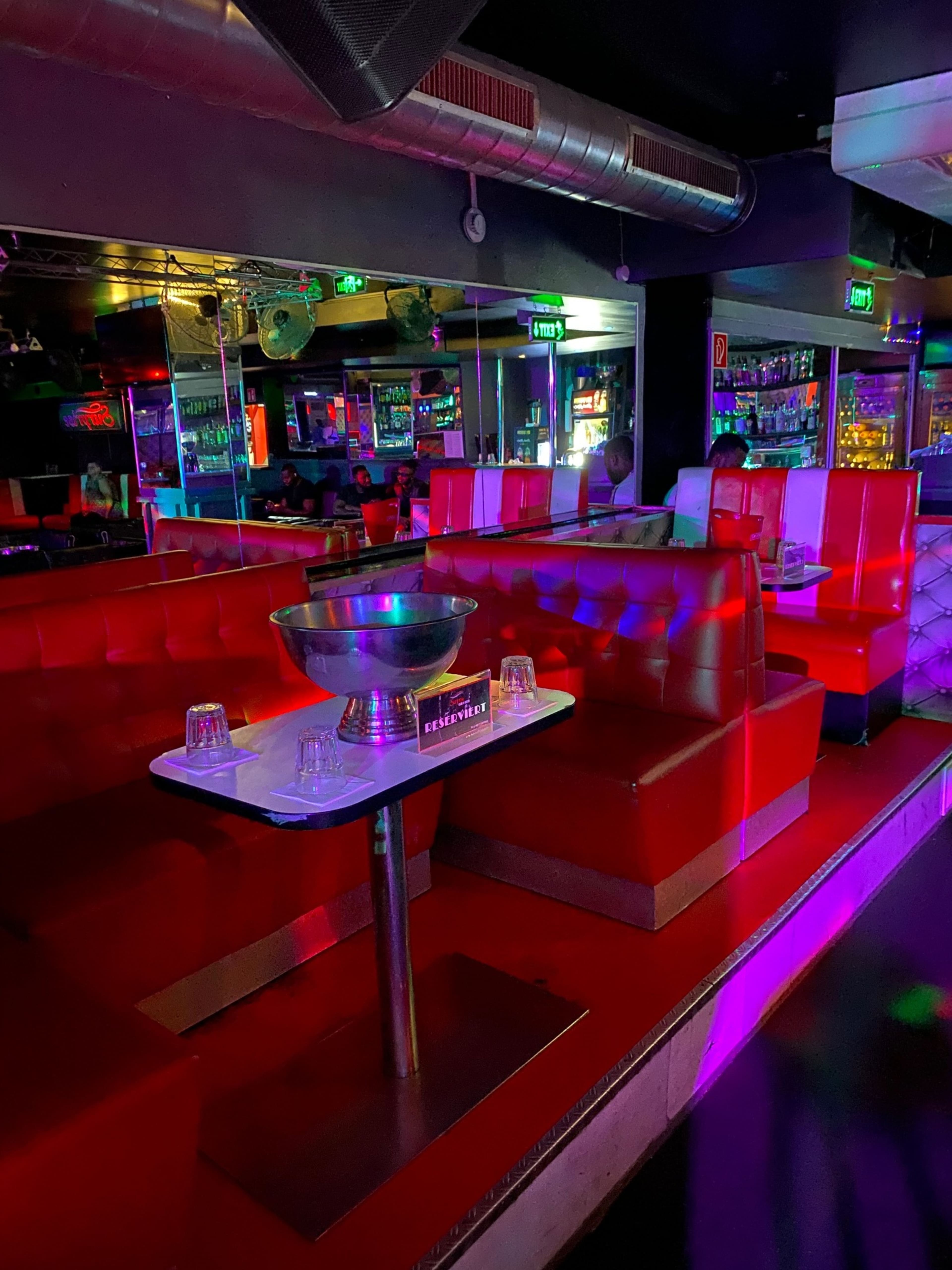 The image shows a dimly lit nightclub interior featuring red leather seating and a central table with a bowl and several glasses.