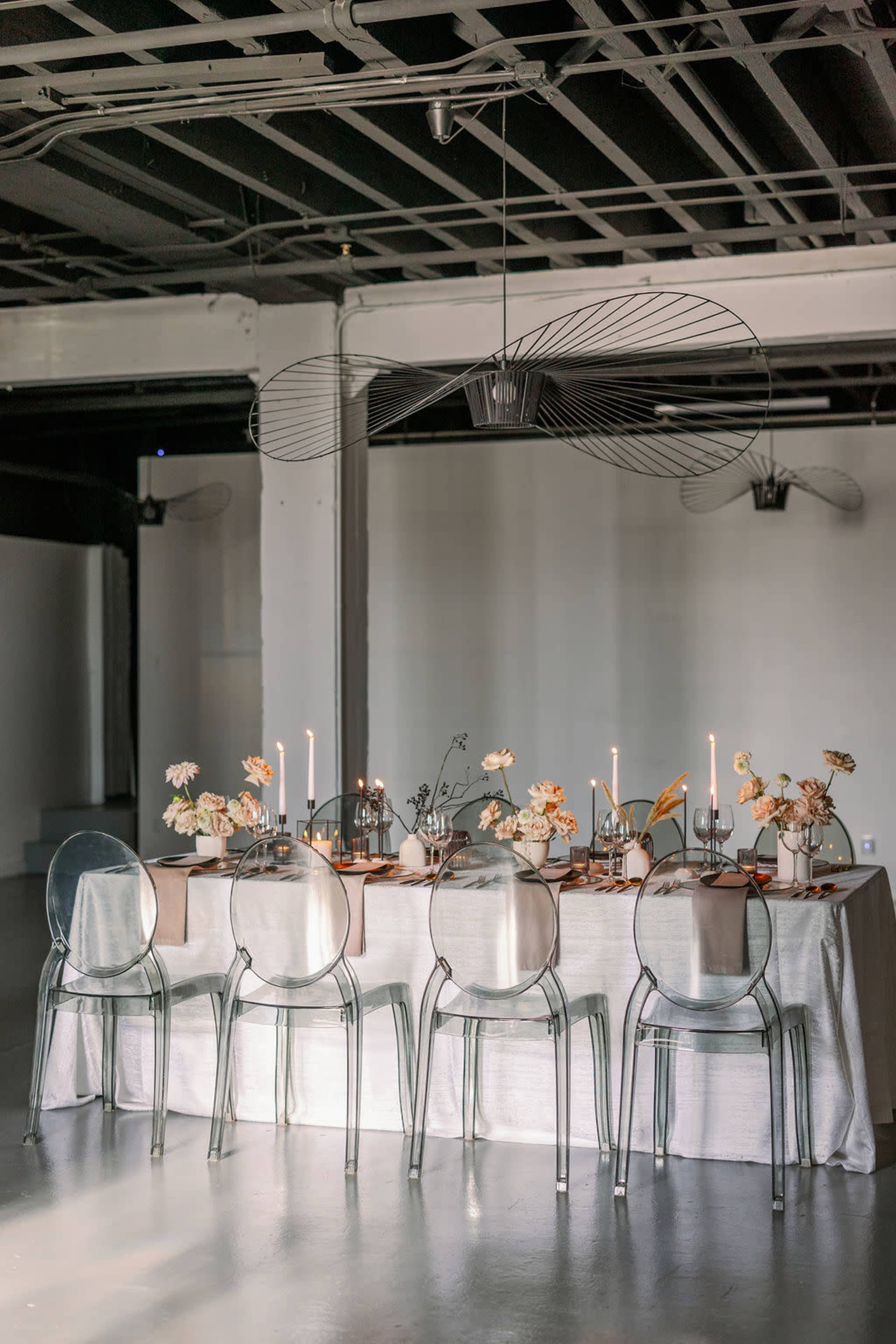 A dining table is elegantly set with candles and floral arrangements under a modern light fixture in a minimalist space.