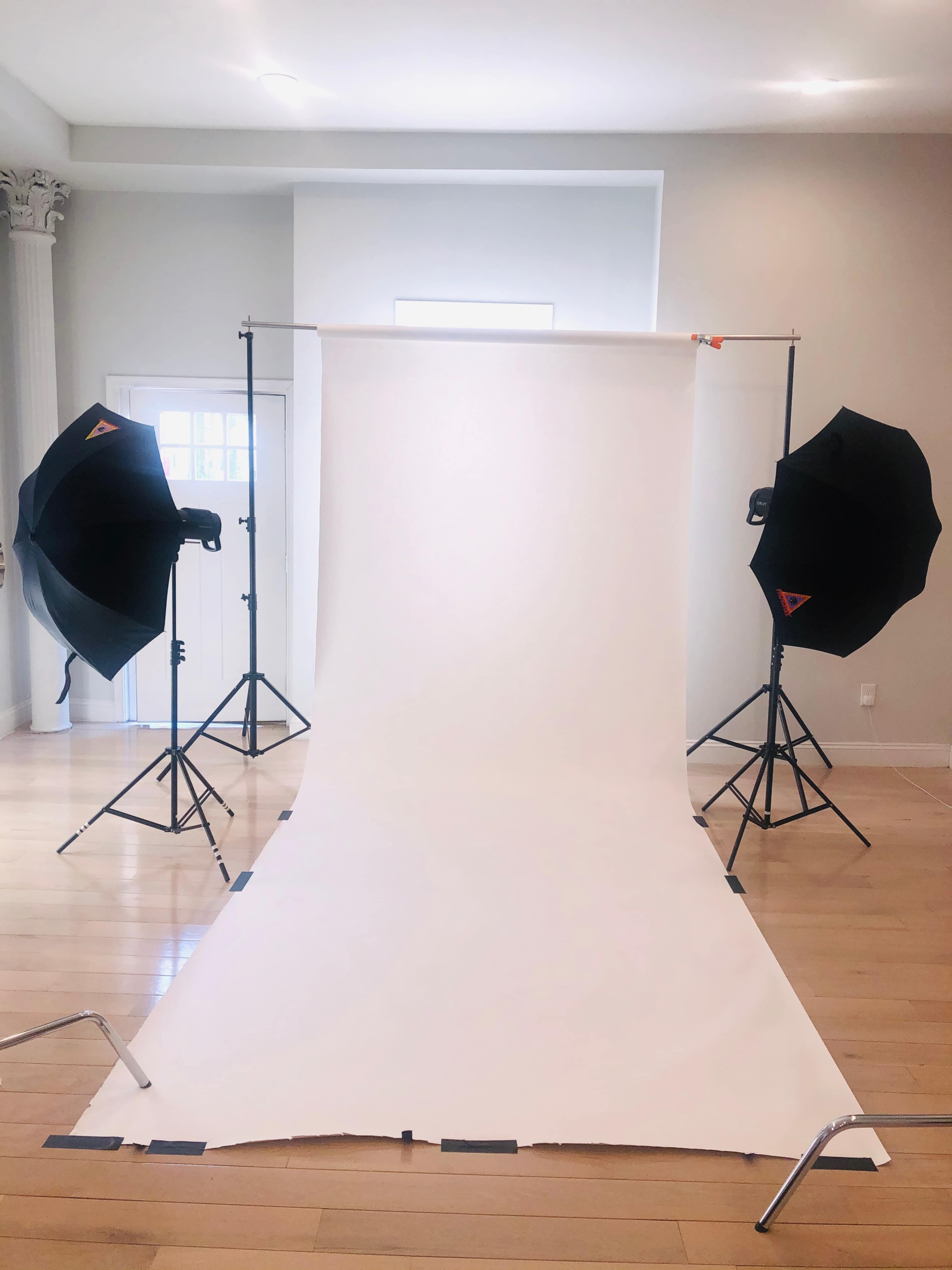 A photography setup with a white backdrop and two lighting softboxes positioned on either side in a bright room.