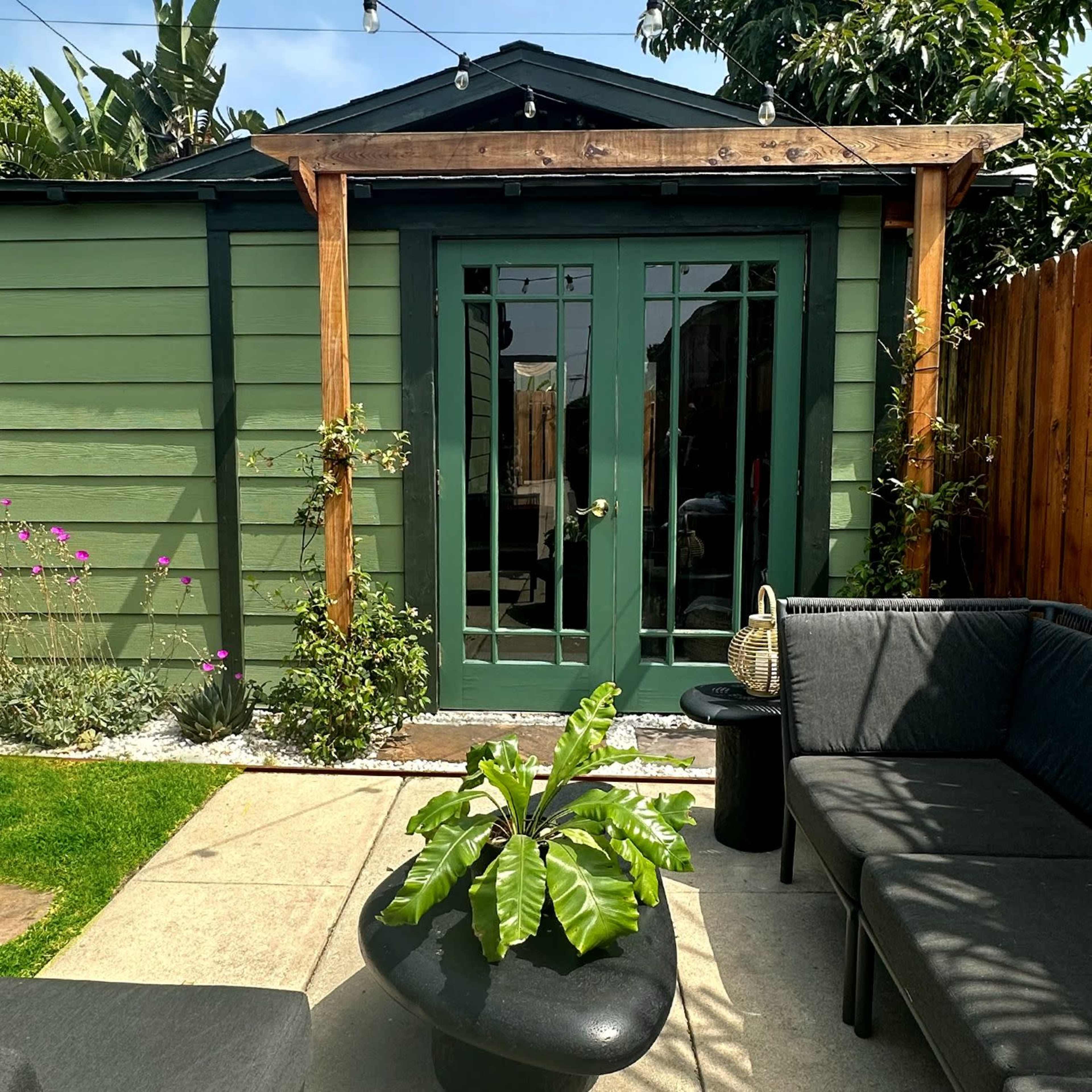 A green shed with double doors is framed by a wooden structure, surrounded by a concrete patio and outdoor seating.