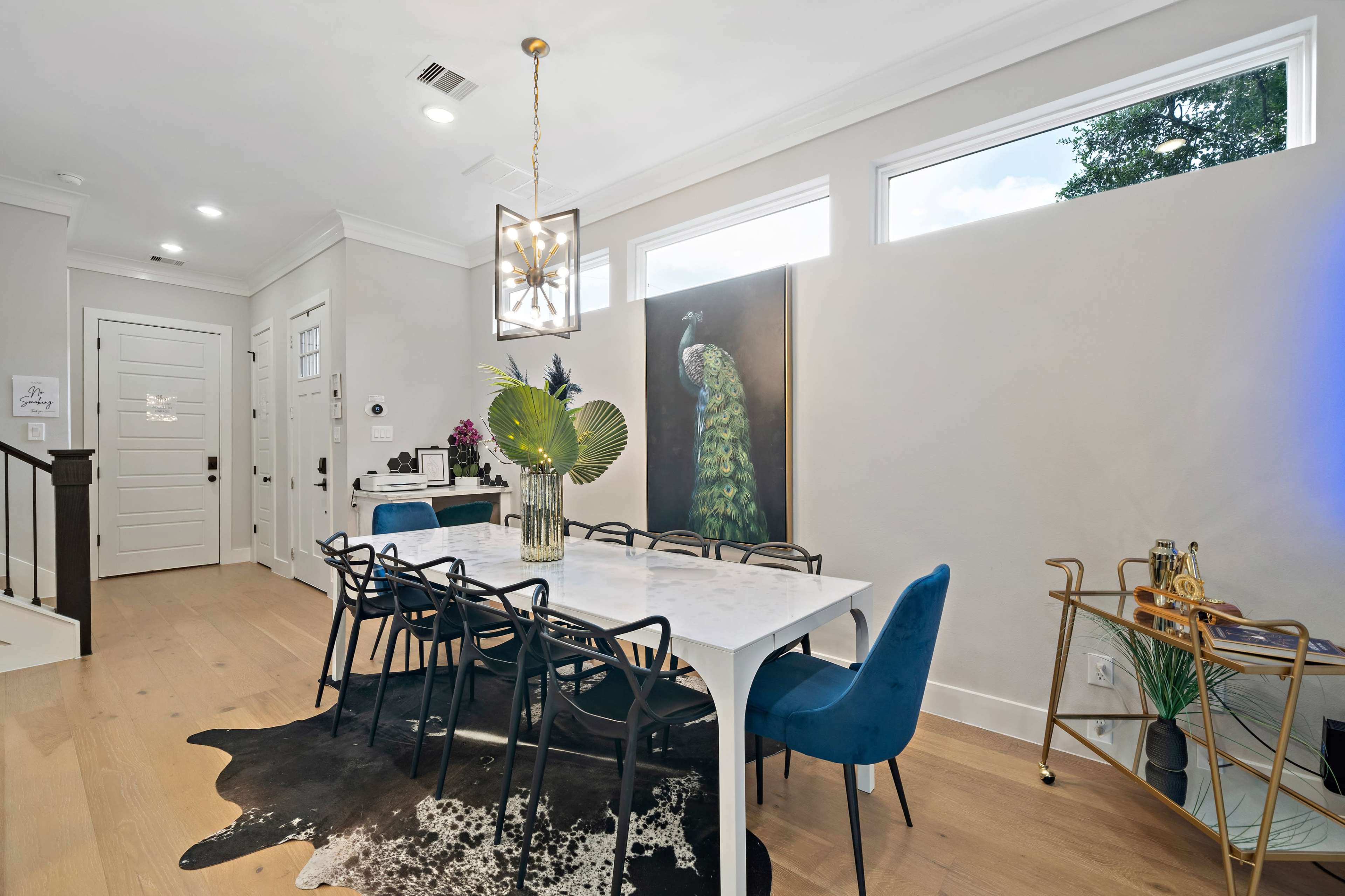 A modern dining area features a large white table surrounded by dark chairs and a decorative peacock painting on the wall.