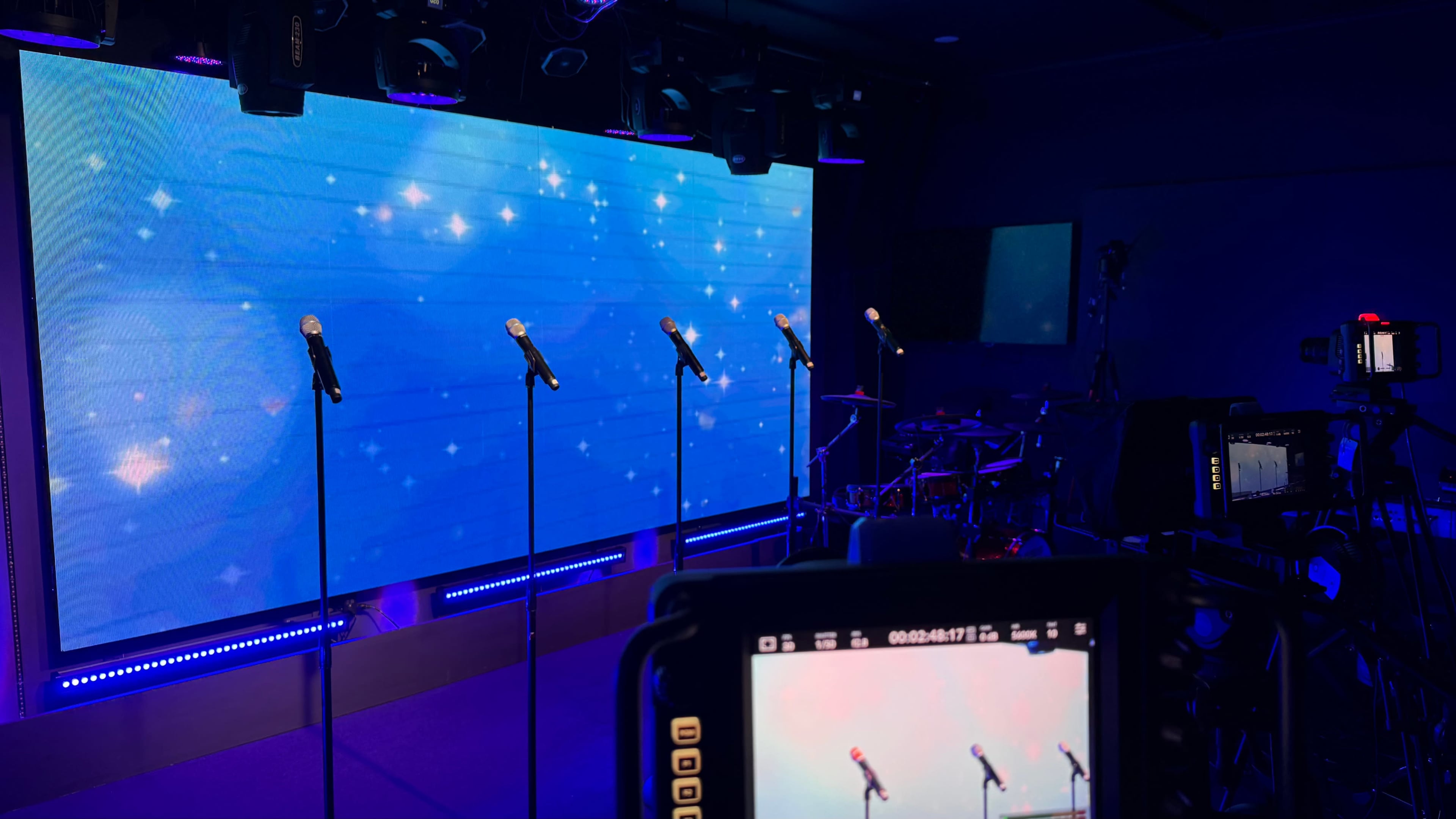 A stage setup features several microphones in front of a large illuminated screen with a blue background, complemented by a drum set and cameras positioned for recording.