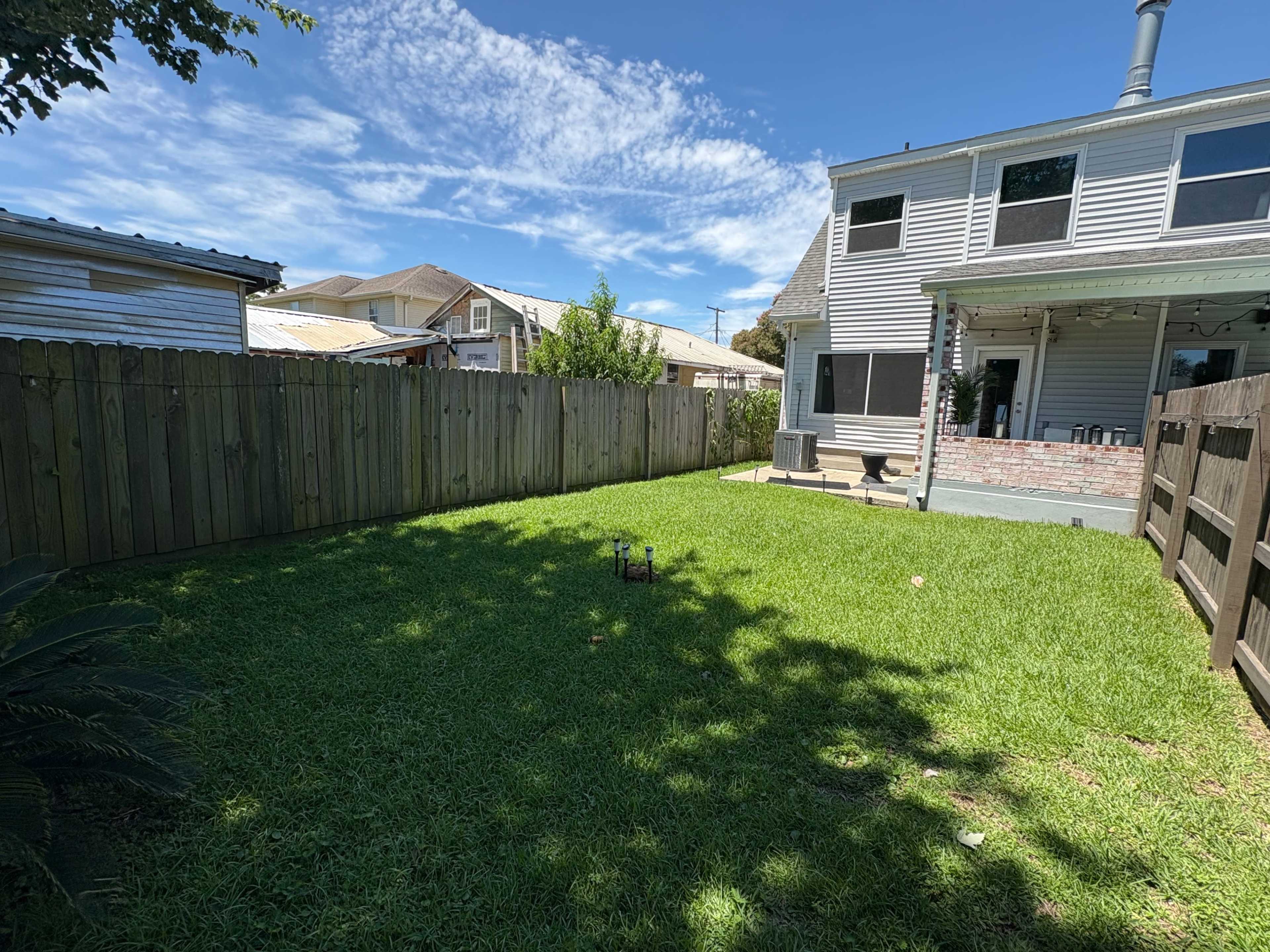 The image shows a well-maintained backyard with green grass, surrounded by wooden fences, and a two-story house in the background under a clear blue sky.