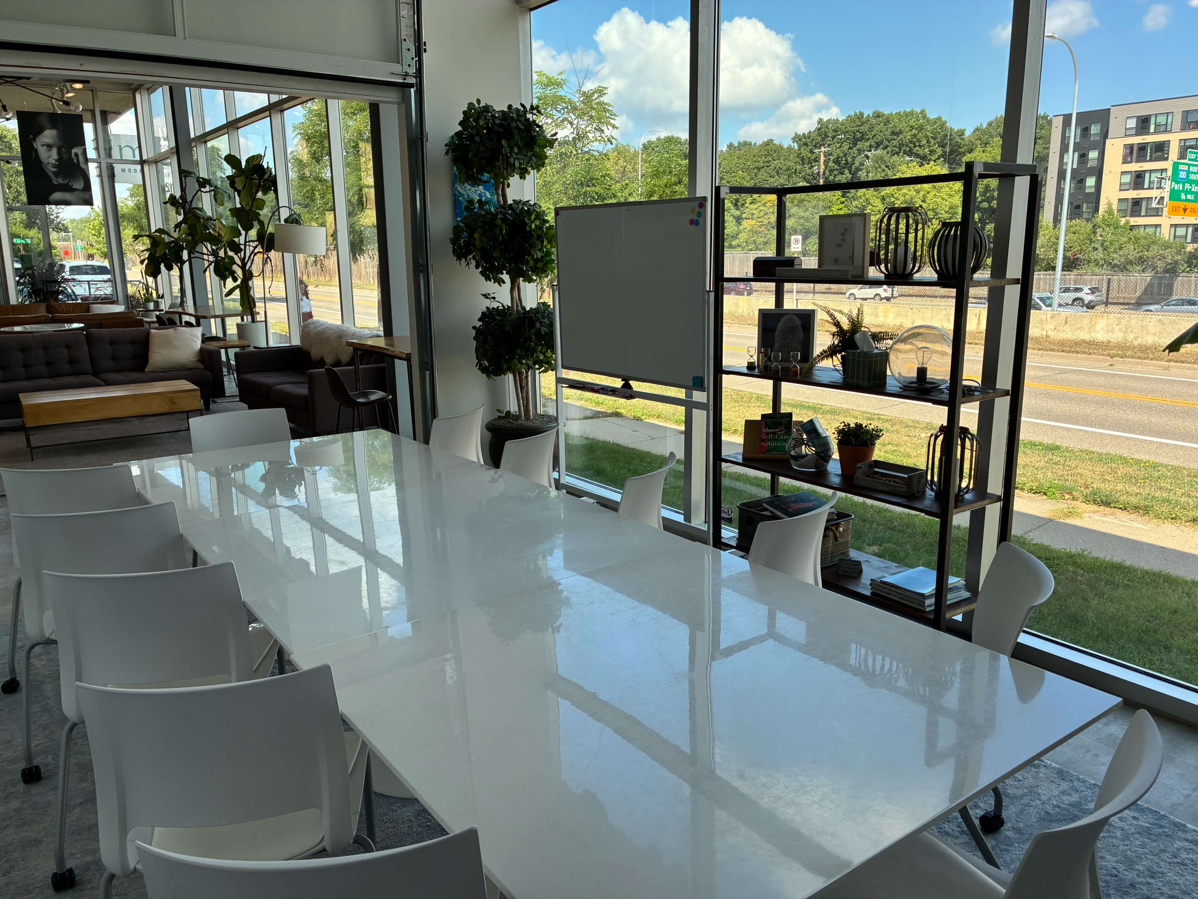 The image shows a modern conference room with a long white table, surrounded by white chairs, large windows letting in natural light, and a shelf displaying plants and decor.