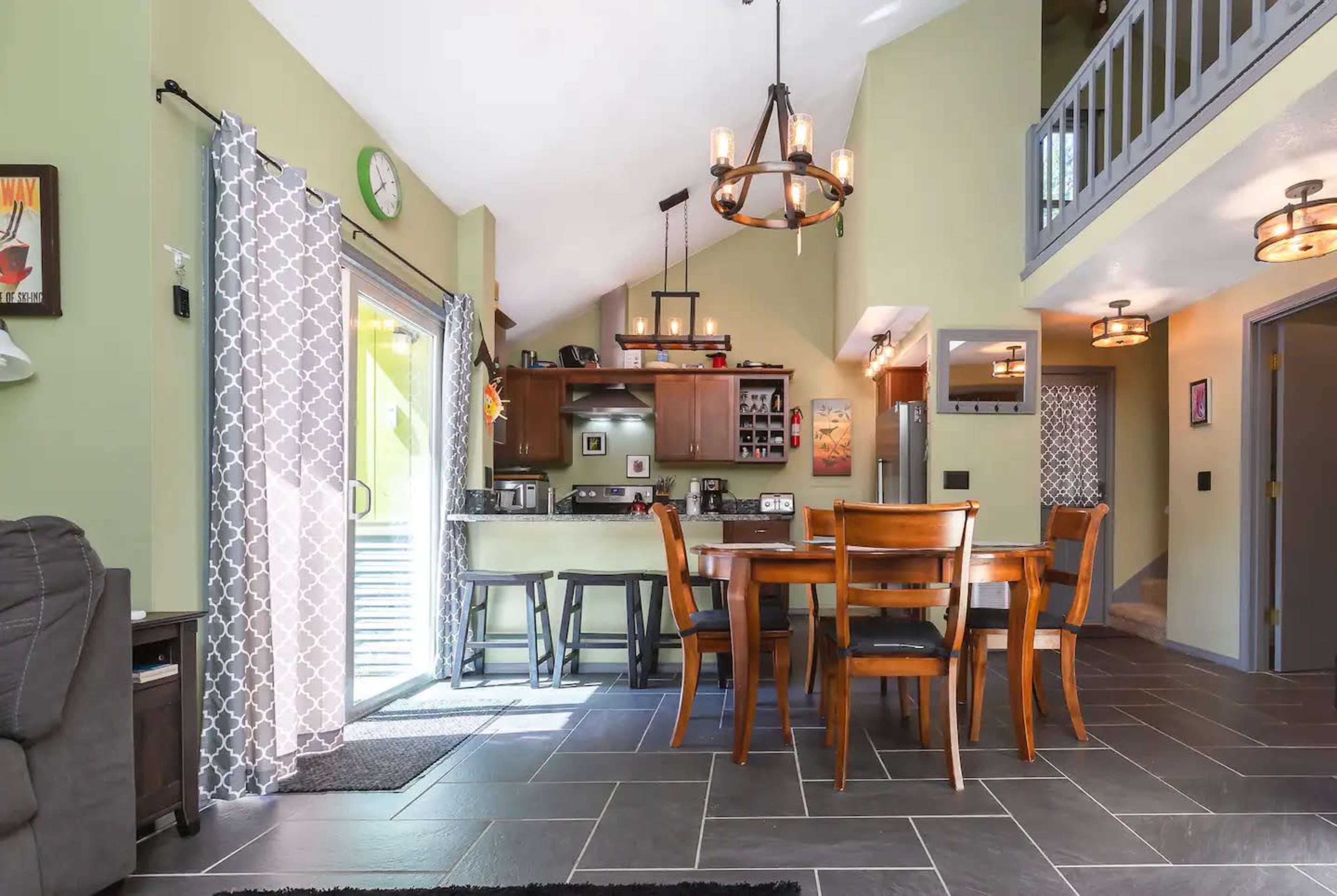 An open-concept living and dining area with a kitchen, featuring tile flooring, a wooden dining table, and large windows.