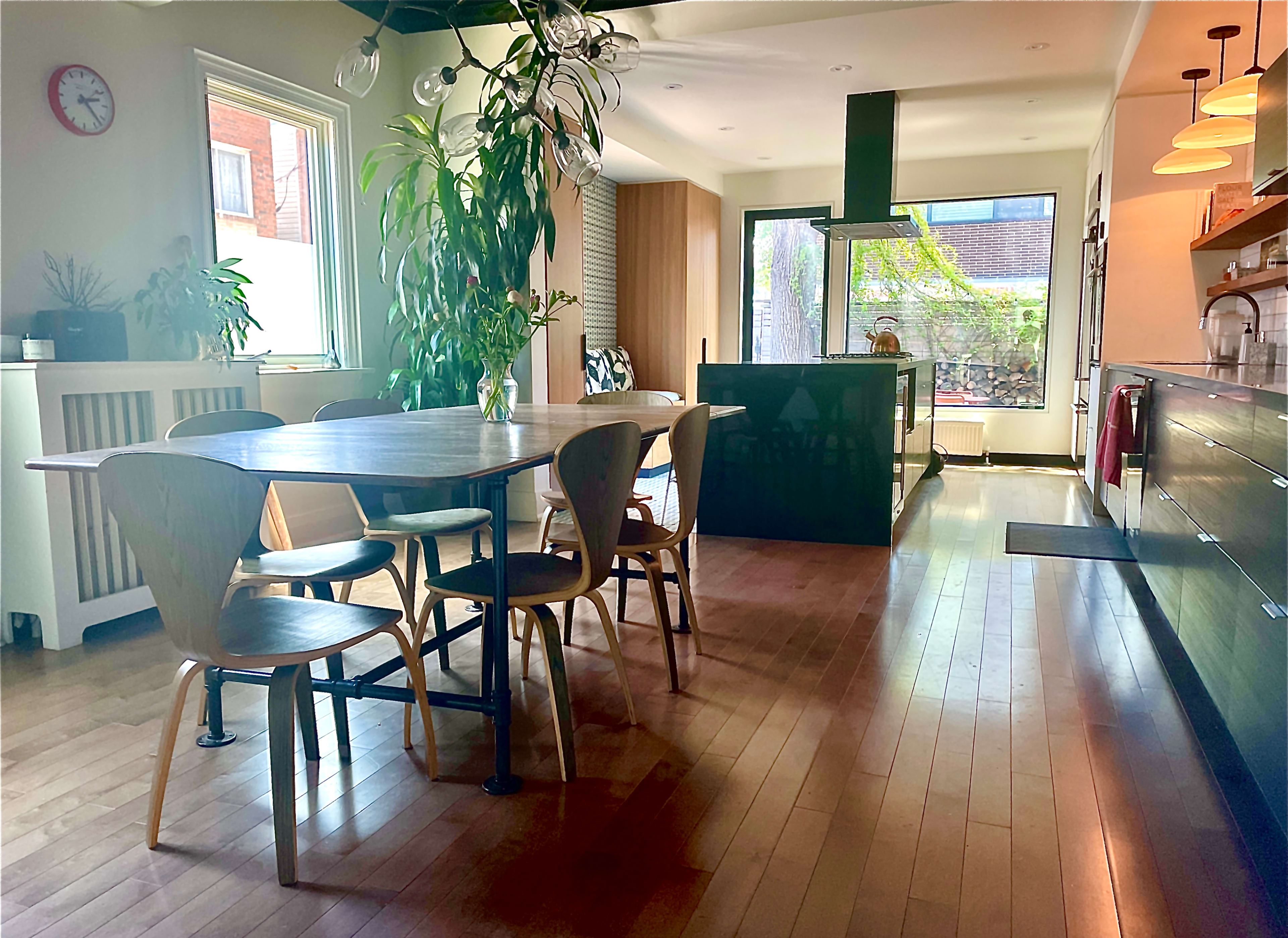 A bright dining area features a wooden table surrounded by chairs, leading to a modern kitchen with a large island and greenery visible through the windows.