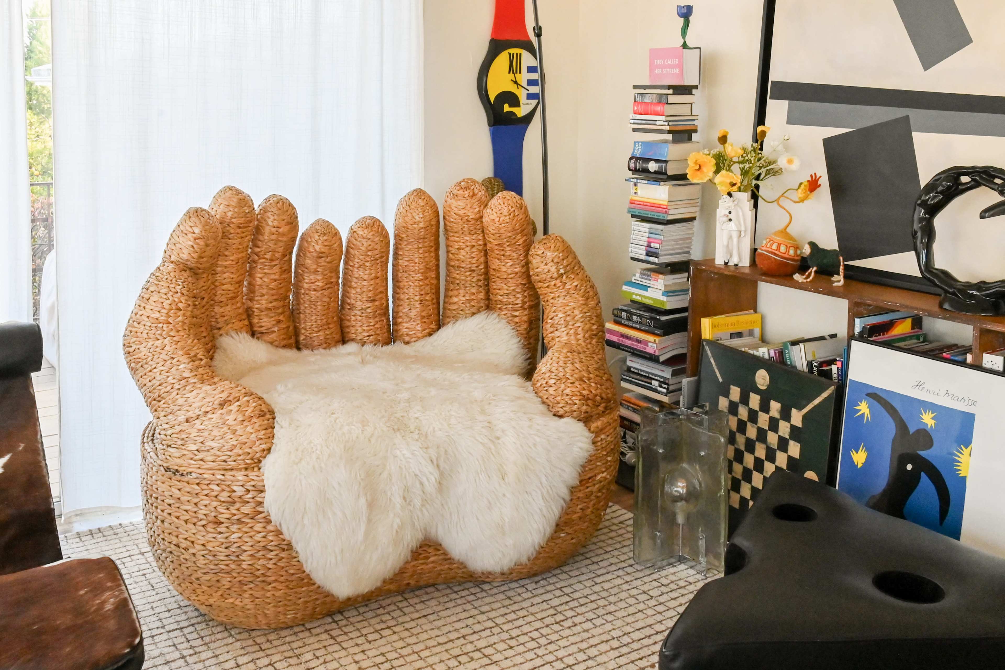A large, woven chair shaped like a hand sits next to a bookshelf filled with colorful books and decorative items in a brightly lit room.