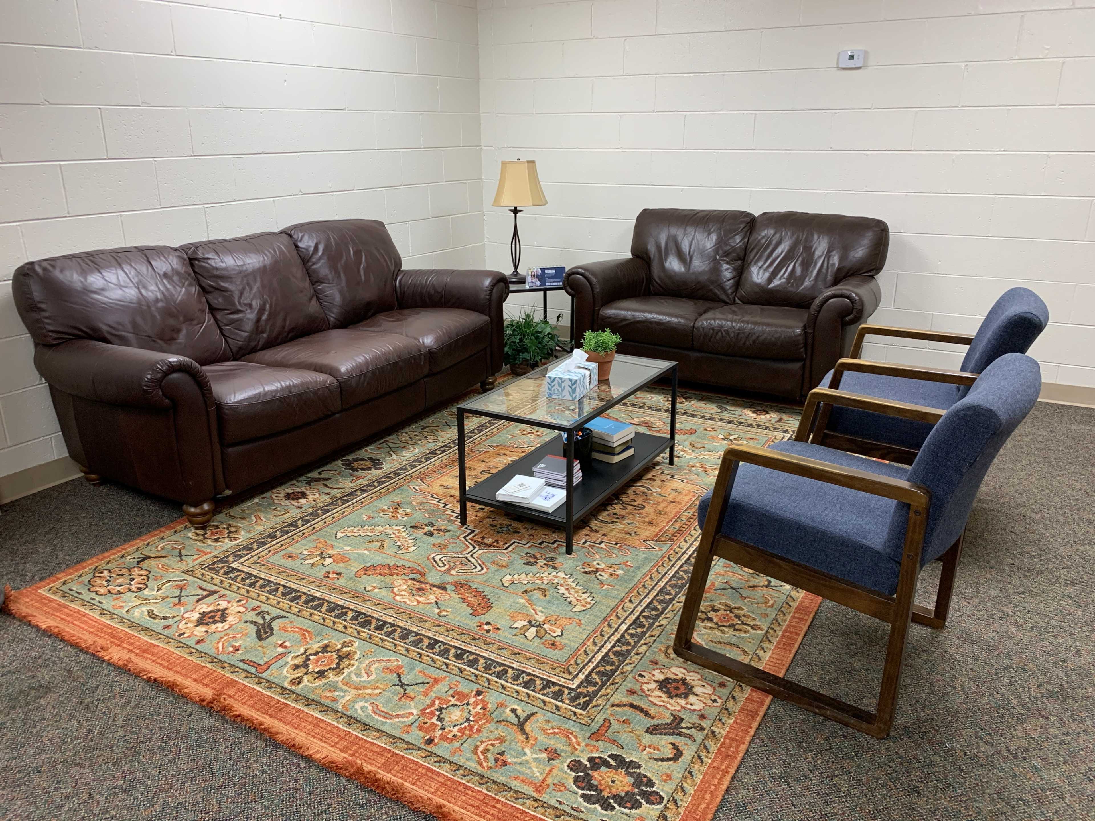 A small seating area with two brown leather sofas, two blue chairs, and a glass coffee table on a patterned rug.