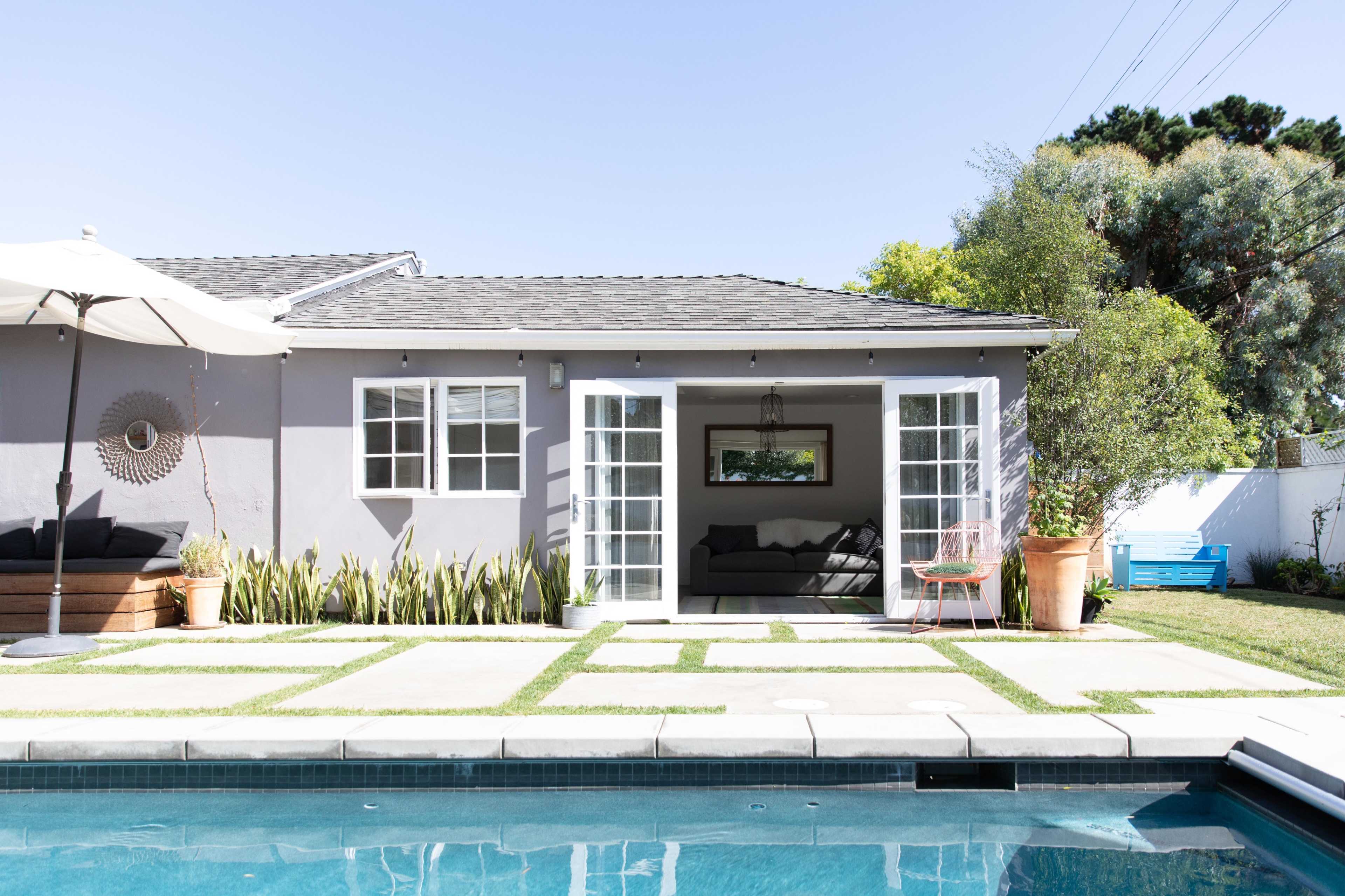 A modern backyard features a swimming pool, a gray house with French doors, and neatly arranged plants and outdoor furniture.