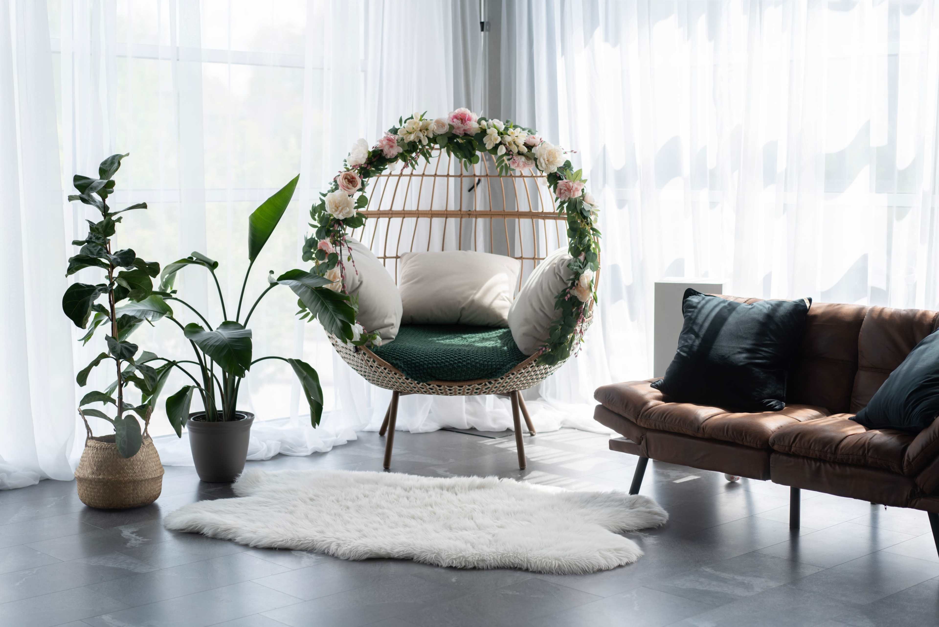 The scene features a circular hanging chair adorned with flowers, positioned beside a potted plant and a brown leather sofa on a soft white rug in a well-lit room.