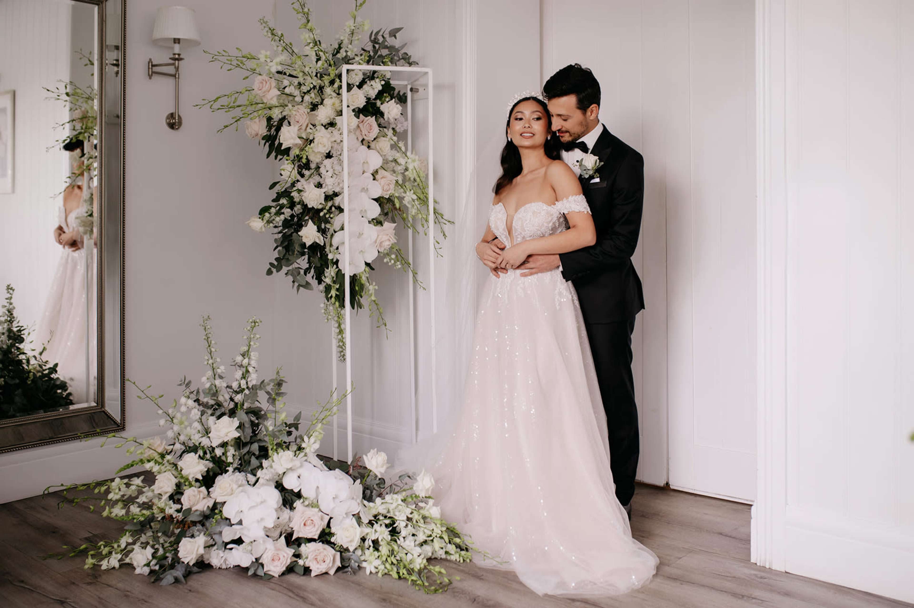 A bride in a strapless gown stands with her groom, surrounded by floral arrangements, in a softly lit interior setting.