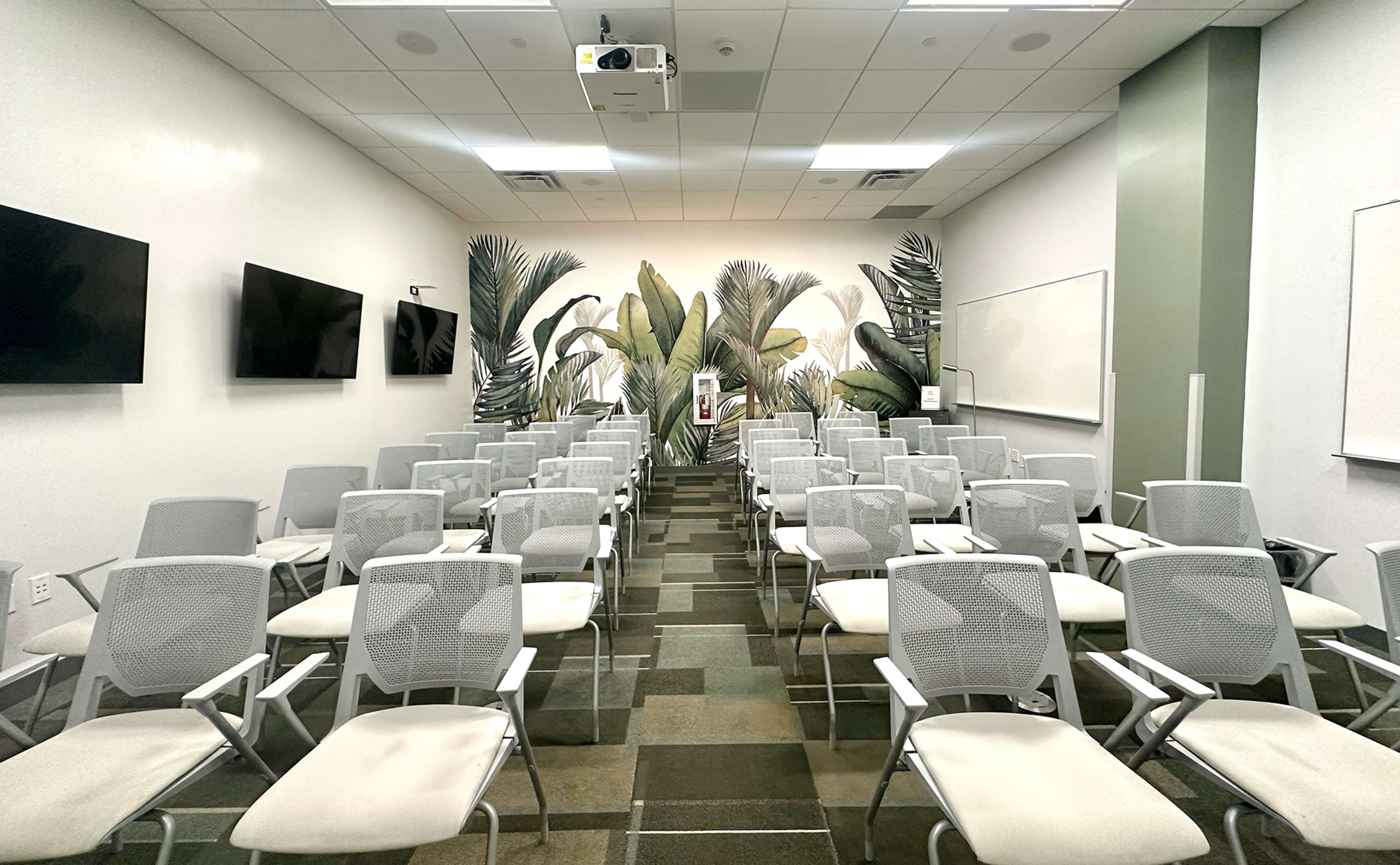 A classroom setup with rows of white chairs facing a projection screen and a wall adorned with palm leaf murals.