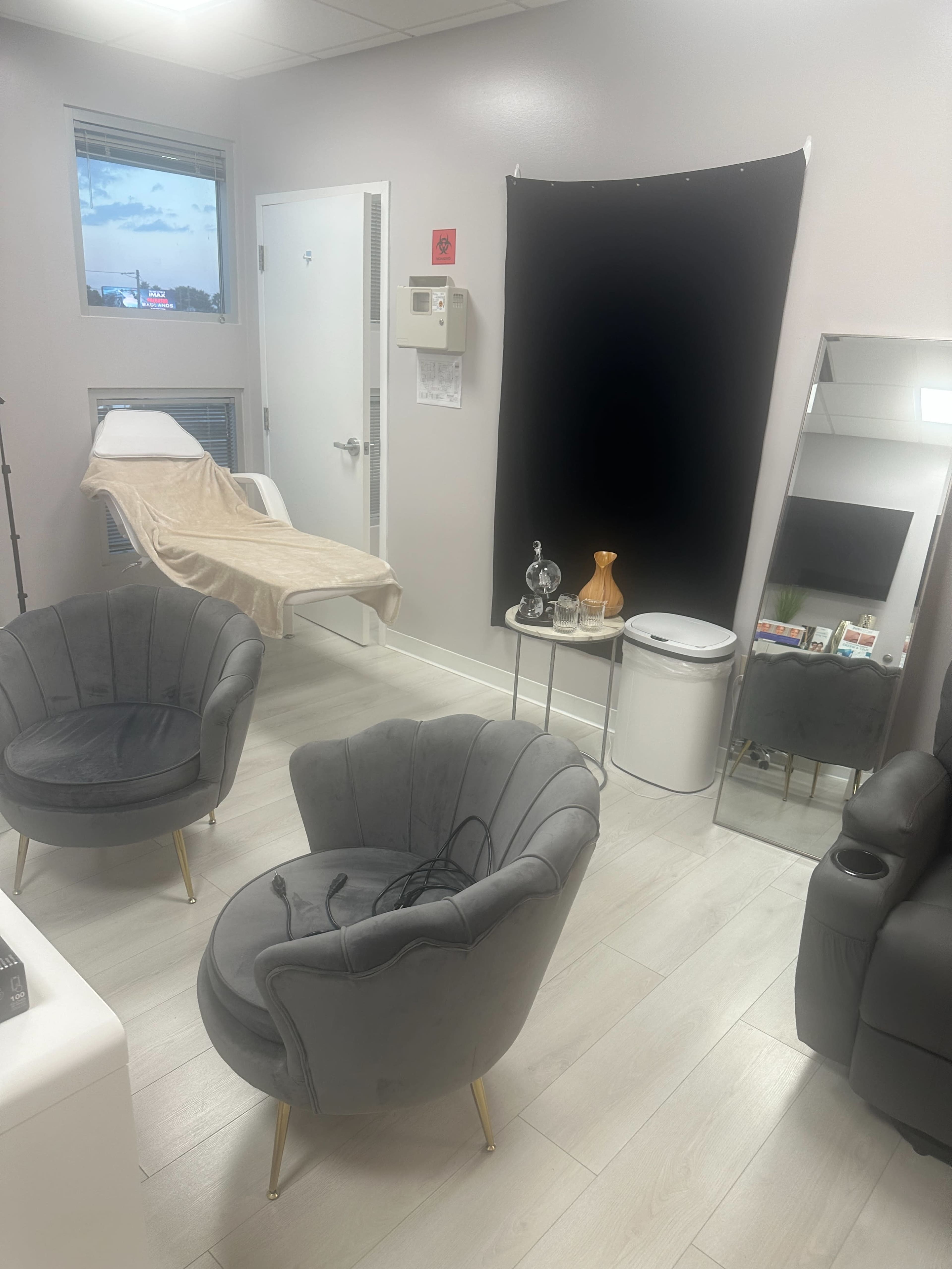 The image shows a clean, modern treatment room with two upholstered chairs, a treatment bed, a mirror, and a black backdrop.