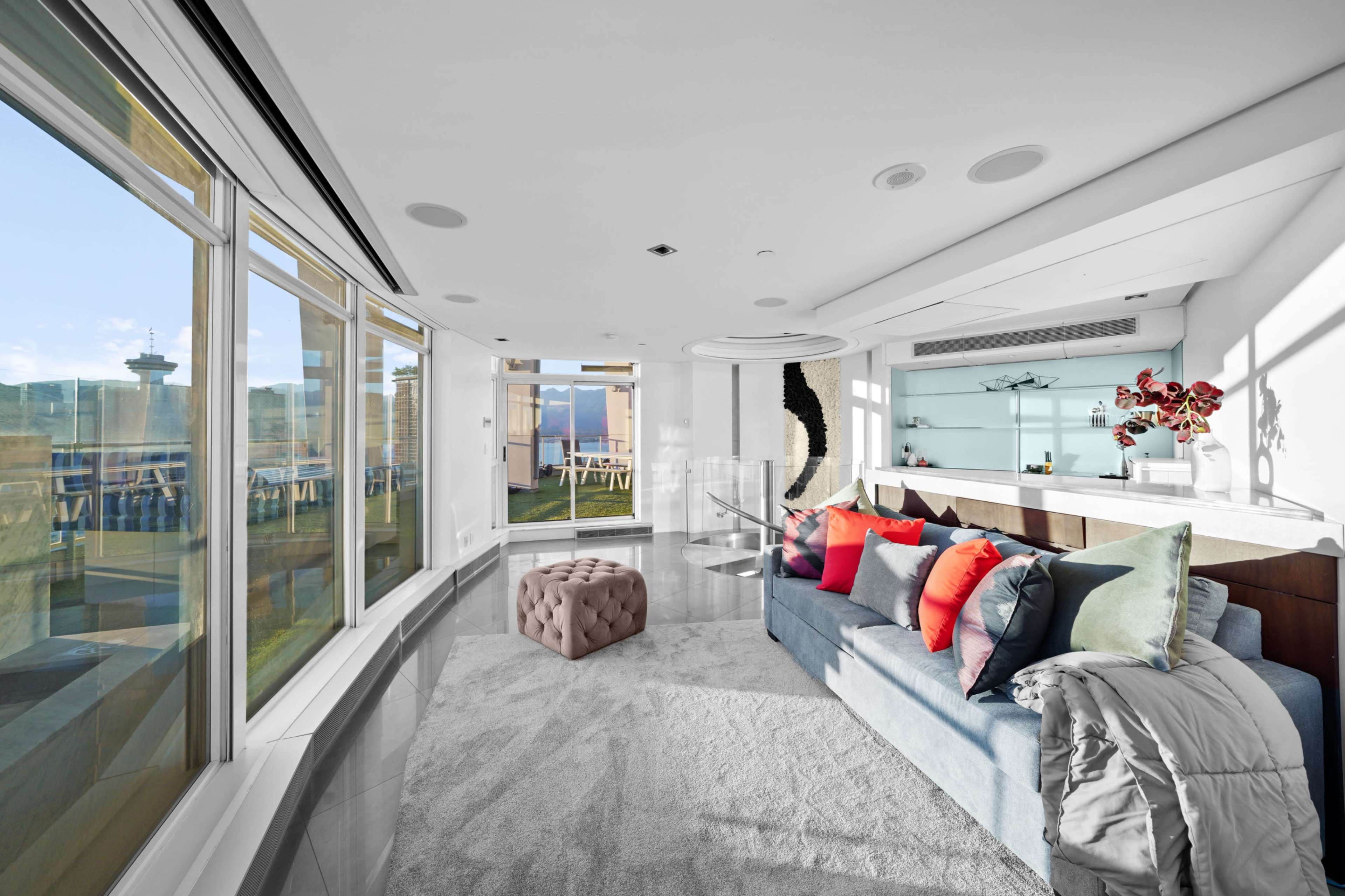 The image shows a modern living room with large windows, a comfortable blue sofa adorned with colorful pillows, and a stylish coffee table, overlooking a cityscape.